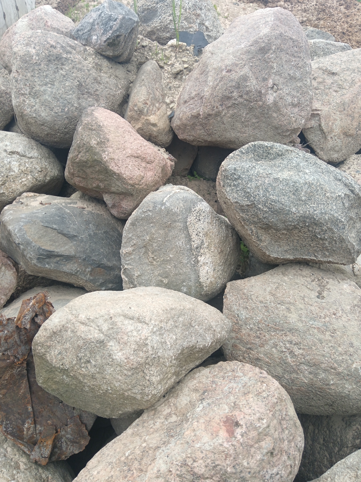 The image shows a pile of various sized, mostly grey rocks. One rock appears to have a pinkish hue. Partially visible on the lower left is some rusted metal. The location is unspecified, though the request mentions Яльгелево, Russia. The image contains no people, actions, or interactions. The environment is natural.
