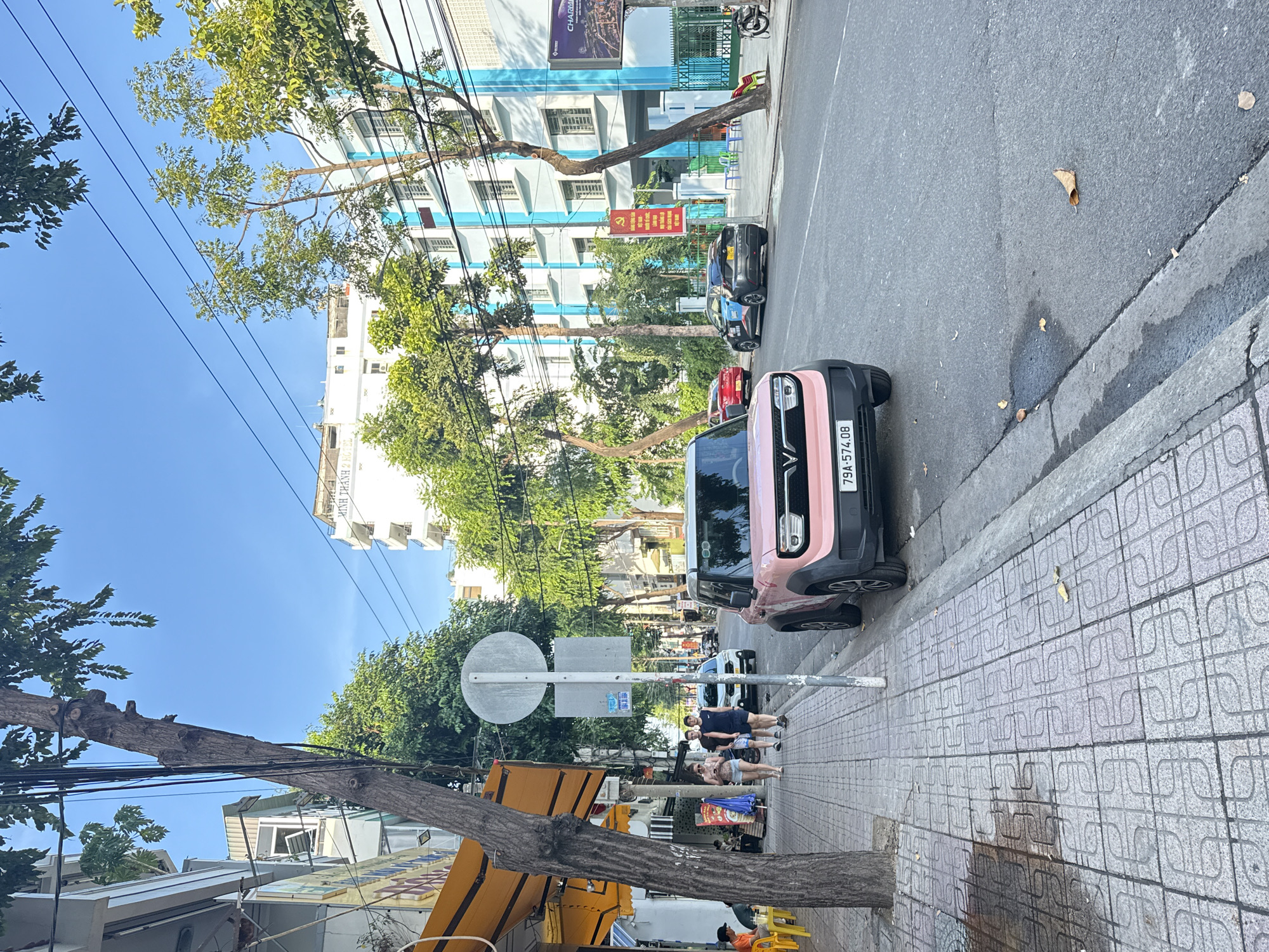 The image captures a street scene in Nha Trang, Vietnam, on a clear day. A pink electric car, with the license plate 79A-574.08, is parked on the right side of the road, facing away from the viewer. A dark-colored sedan is parked behind it, also on the right side of the road.
On the left side of the street, a sidewalk is visible, paved with patterned tiles. A few people are walking along the sidewalk, including a woman in a light-colored top and denim shorts, and two men in casual wear. A tree with a thick trunk stands on the sidewalk, its branches extending over the street and sidewalk. A yellow awning is visible extending from a building on the left.
The background features multi-story buildings, some with blue accents, and a clear blue sky above. There are also trees lining the street and sidewalk, contributing to the urban but green atmosphere of the location. Streetlights and power lines are also present, indicating it's a typical urban street. A red political banner with Vietnamese text is displayed on a building in the background.