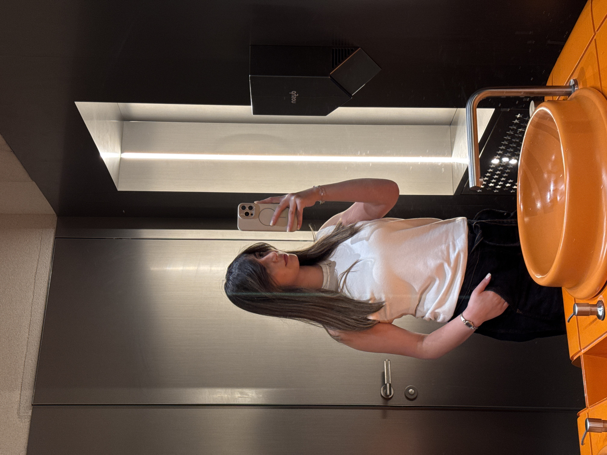 A young woman is captured in a mirror selfie within what appears to be a modern public restroom or washroom in Yerevan, Armenia. She has long, dark hair with lighter streaks and is wearing a light-colored t-shirt and dark bottoms, accessorized with a watch and bracelet. She holds a light-colored iPhone with a MagSafe attachment, using it to take the photograph.
The setting features a sleek design with dark and metallic surfaces. Above the woman's head, a rectangular black unit is mounted on the wall, displaying the reversed text "qilou". A prominent linear light fixture is recessed into the dark ceiling panel, illuminating the space. To the right, an distinctive orange, bowl-shaped fixture, possibly a urinal or a unique sink, is visible with metallic piping. The mirror reflects the immediate surroundings and a patterned surface of small stars from behind the viewer, visible on the metallic pipe. To the left of the woman, a metallic door with a latch handle and a round lock mechanism is present, alongside a textured, light-colored wall. The overall ambiance suggests a contemporary and well-lit indoor environment.