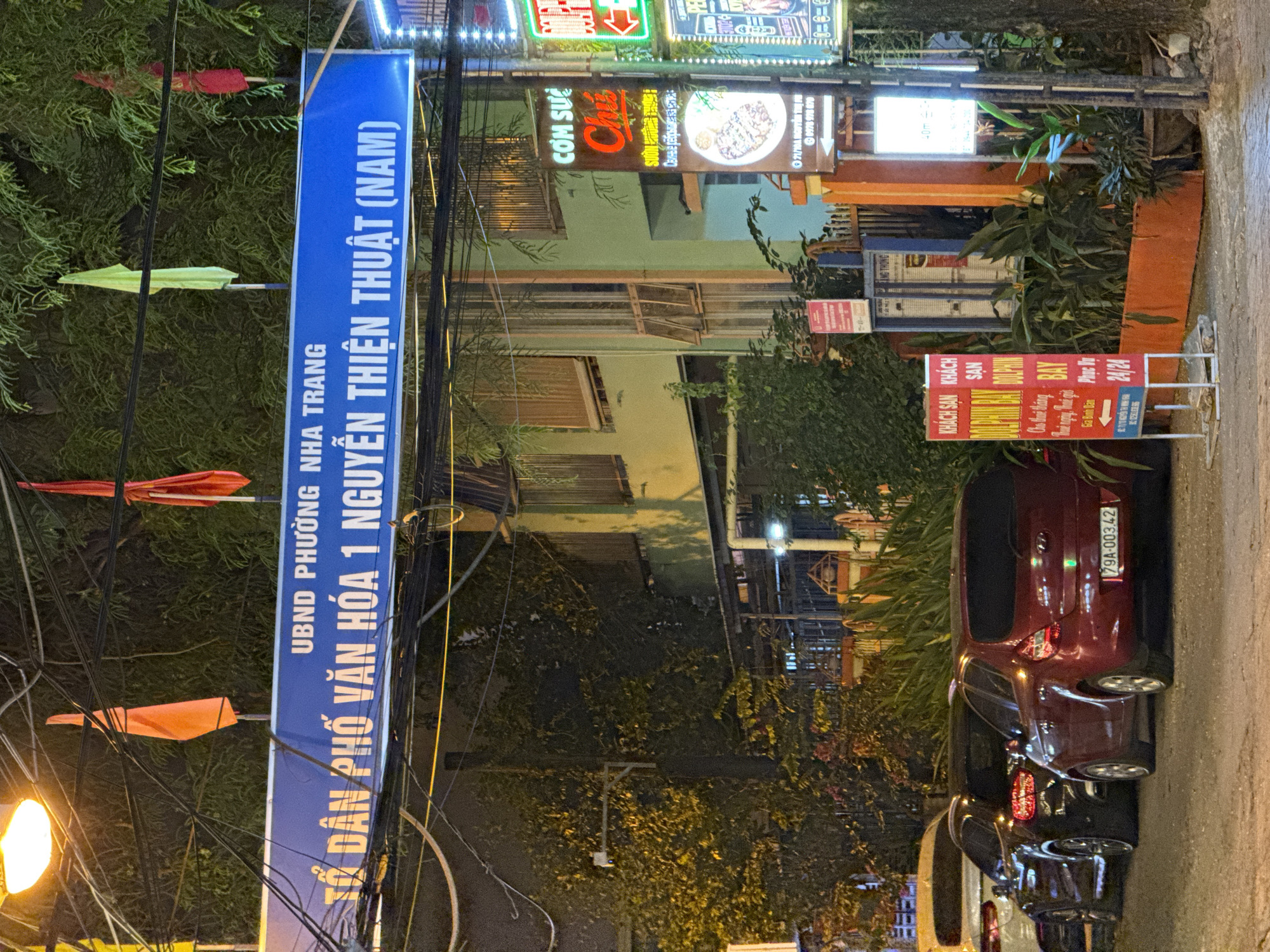 The image depicts a street scene at night in Nha Trang, Vietnam. A prominent blue sign with white lettering reads "UBND PHƯỜNG NHA TRANG TỔ DÂN PHỐ VĂN HÓA 1 NGUYỄN THIỆN THUẬT (NAM)". Several flags are visible, including orange, green, and red. The street is lined with buildings, some of which have illuminated signs. One sign advertises "CƠM SƯỜN CHUỘT" with an image of food. Another sign for "KHÁCH SAN DOLPHIN BAY" is also visible. Cars are parked along the street, including a red hatchback with license plate "79A-003.42". Trees and vegetation are present, with some illuminated by streetlights. The overall scene suggests a commercial and residential area at night.