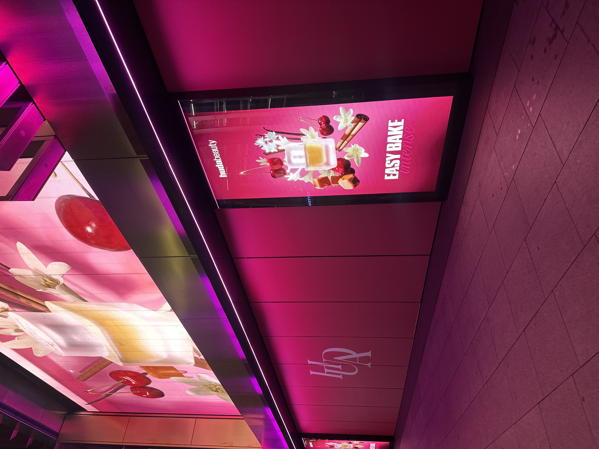 The photograph shows a section of a building in London, UK, decorated with Huda Beauty advertisements. The building's exterior is primarily pink, featuring a logo and lit displays. The displays show product advertisements, depicting images of ingredients such as cherries, vanilla, and cinnamon sticks. The walls feature straight lines and the floor is tiled in a matching color. The lighting is purple, highlighting the building's lines.