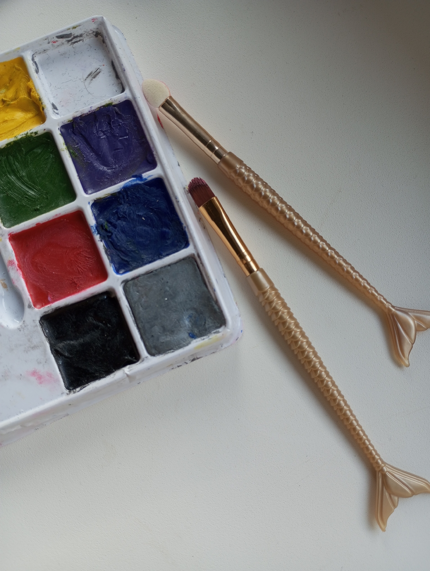 The image displays a close-up, high-angle view of a used paint palette and two unique paintbrushes resting on a plain, light-colored surface, likely a table.
The main subject is a rectangular, white plastic palette containing multiple square wells filled with various colors of paint. Visible colors include vibrant yellow, deep green, rich purple, bright red, dark blue, black, and gray. One well appears mostly empty or contains very little paint. The paints have a thick, creamy texture, suggesting they might be face paints or cream-based theatrical makeup. The palette shows signs of prior use, with smudges and residue around the edges of the wells.
Adjacent to the palette are two distinct paintbrushes. Both brushes feature decorative golden handles with a textured, scale-like pattern that culminates in a stylized mermaid tail shape at the end. One brush has a fluffy, rounded white head, suitable for blending or applying larger areas of color. The second, smaller brush has an angled, flat head with dark bristles, possibly stained with a reddish hue, suggesting it has been used.
The arrangement implies that painting activity has either just concluded or is about to begin. The lighting in the image is soft and even, suggesting natural daylight. No people are visible, and no text is discernible. The scene is set indoors, possibly in a home or studio in Pervomaysky, Russia, as per the provided location context.