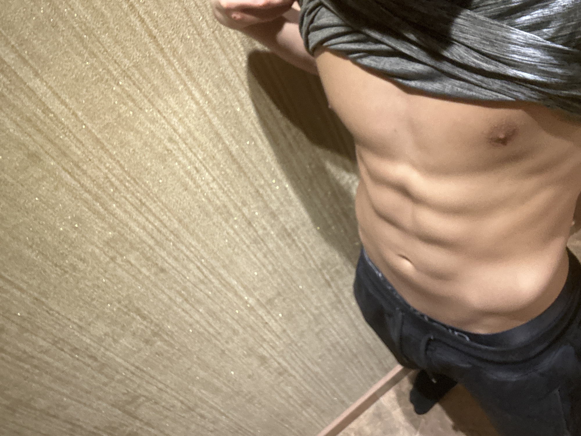 A medium shot of a muscular man with visible abdominal muscles. He is wearing dark blue or black jeans and a gray shirt pulled up to reveal his torso. The background is a textured, light brown wall. The location is impossible to determine.