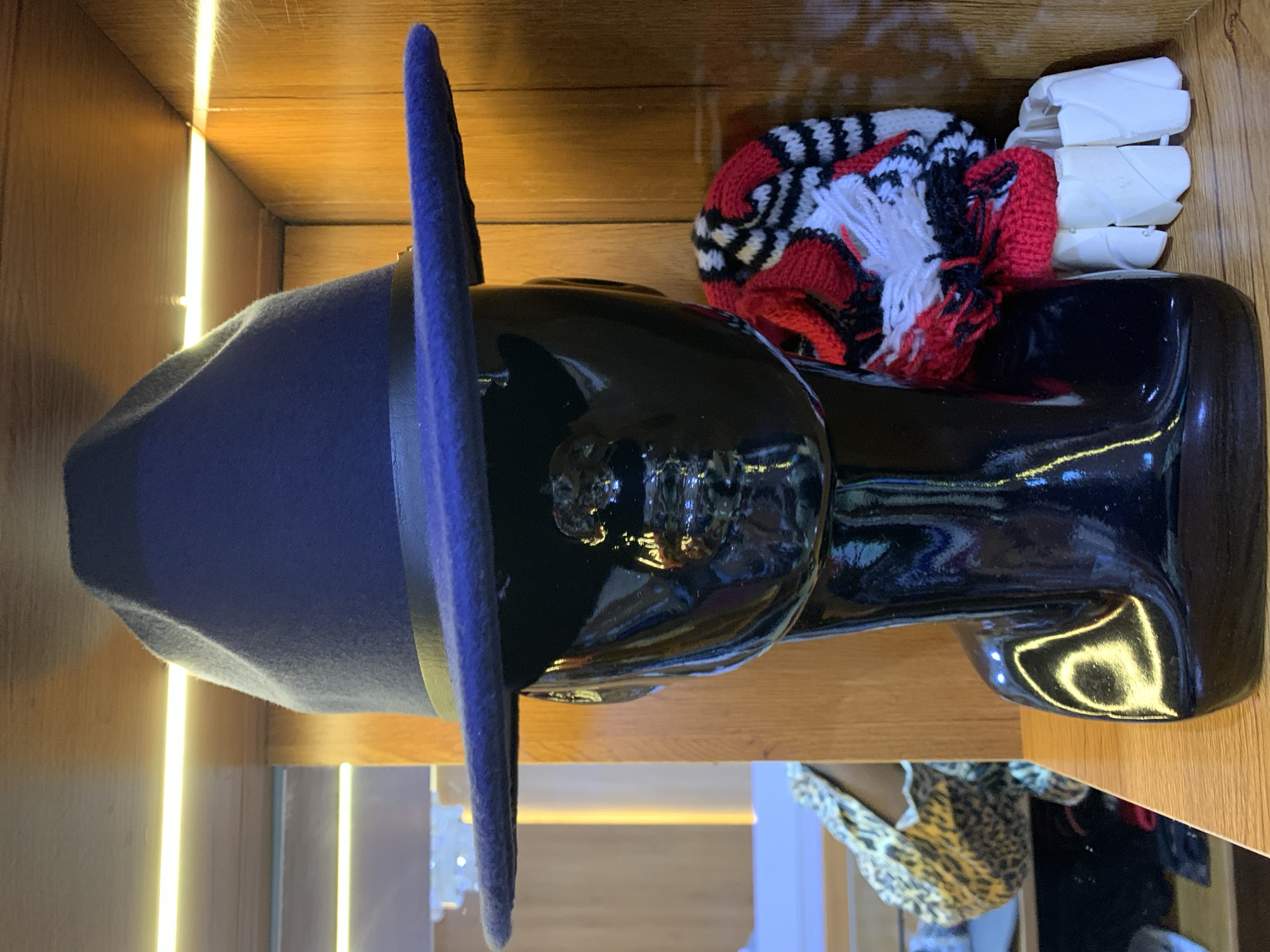A dark blue fedora hat is displayed on a black mannequin head in a retail setting, likely a hat shop or boutique. The hat has a wide brim and a black ribbon band. To the right of the mannequin, a knitted beanie with a red, white, and black striped pattern and a red pom-pom is visible. Below the beanie, a white segmented bracelet or cuff is on display. In the background, wooden shelves are illuminated by warm lighting, suggesting an indoor environment. To the left of the mannequin, a glimpse of leopard-print fabric and other items are visible on lower shelves, hinting at a broader selection of merchandise. The overall presentation is that of a display for fashion accessories. This image does not contain information that would allow for a specific location within Gura Top, Nigeria.