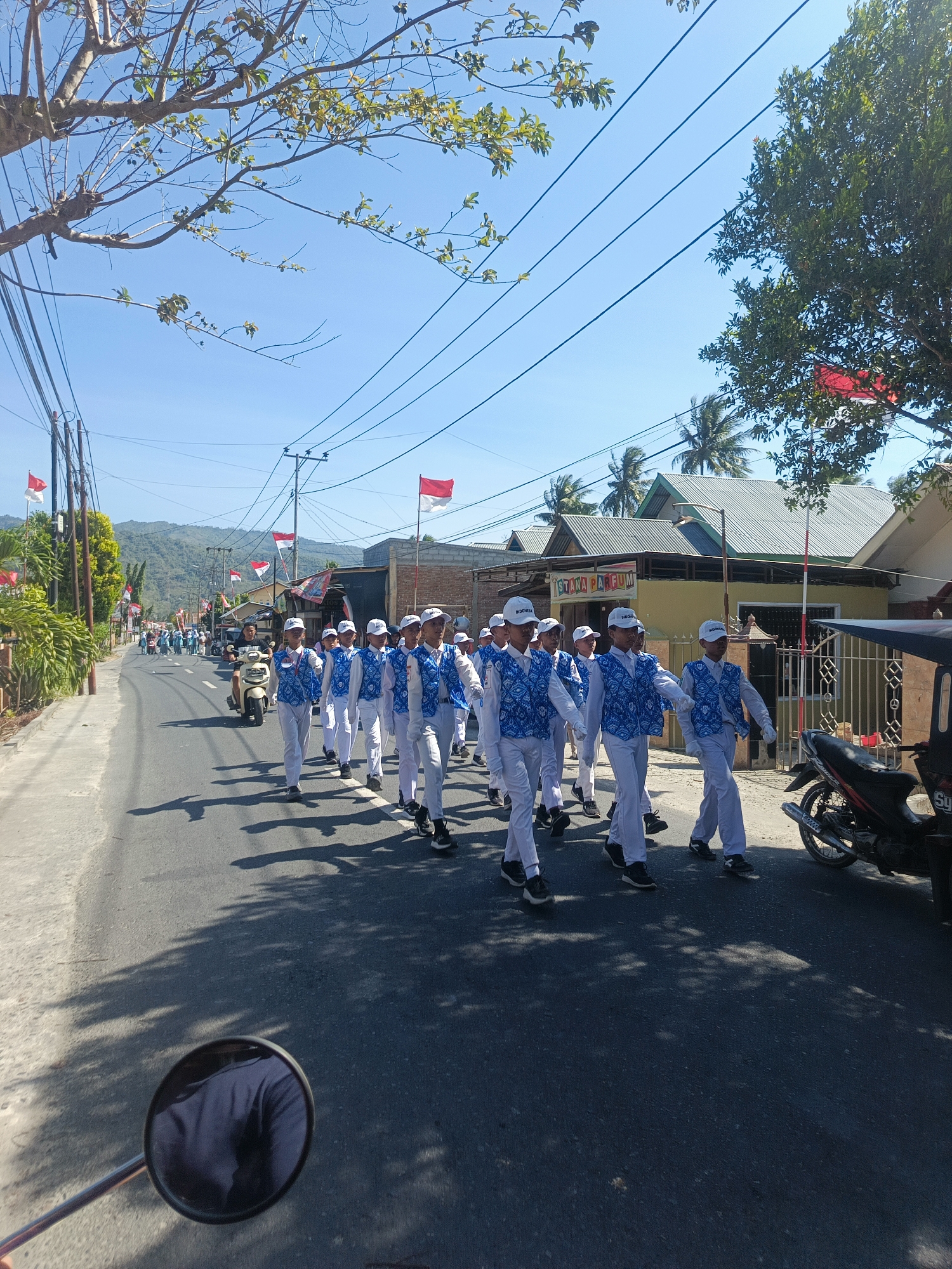 A marching band comprised of young people in uniform walks down a street in Tenggela, Indonesia. The participants wear white caps and vests with a blue and white pattern over white shirts and pants. They are marching in formation on a paved road lined with trees and buildings. Red and white Indonesian flags are visible along the street and on some of the buildings. In the foreground, the reflection of a person on a motorcycle in its side mirror is visible. The sky is clear and blue, indicating a sunny day.
