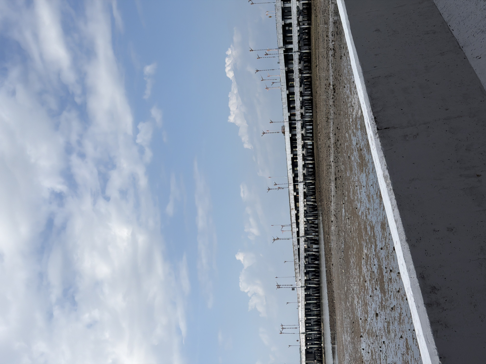 An eye-level, full shot captures a partially exposed seabed under a bright, cloudy sky, with a long, elevated concrete bridge extending horizontally across the frame. The foreground features a concrete barrier, angled diagonally from the bottom left to the bottom right, its textured surface showing some wear and tear. Behind the barrier, the seabed is a mosaic of shallow, muddy water and exposed sand, dotted with dark debris and small rocks. Several tall, slender poles with orange lights are visible on the bridge, spaced intermittently. The bridge itself is a substantial concrete structure supported by numerous vertical pilings that disappear into the water. The sky above is a pale blue, generously populated with scattered white clouds of varying sizes and densities. The scene is devoid of any human presence or activity. This scene is likely part of the southern coast of Denpasar, Indonesia, possibly near the Sanur area, given the elevated bridge infrastructure commonly found over shallow coastal waters.