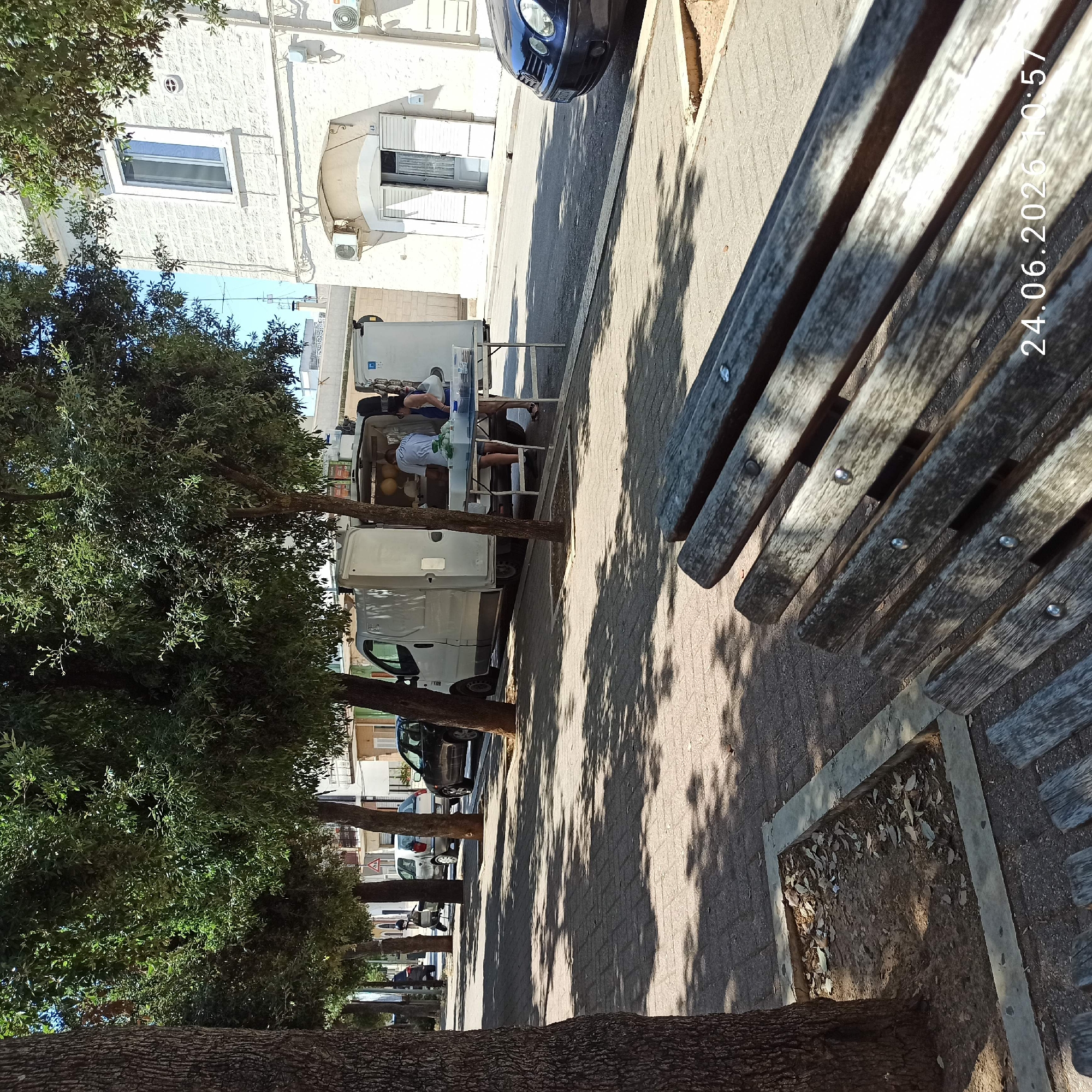 This image depicts a street scene in Bitonto, Italy, likely during the daytime, given the bright sunlight and shadows cast by trees. In the foreground, a weathered wooden park bench is visible, with its slats showing signs of age. Behind the bench, a tree trunk stands on the left, and further back, a sidewalk paved with interlocking bricks runs parallel to the street. A row of trees with dense green foliage lines the sidewalk, casting dappled shadows on the pavement.
On the street, several parked cars are visible, including a black sedan on the right. A white van is parked on the left side of the street, with its rear doors open. Two individuals are interacting near the open van, with one person, wearing a light-colored shirt, appearing to be behind a table set up in front of the van. The second person, wearing a dark top, stands next to the table. A portable table is set up in front of the van, displaying some items, possibly goods for sale.
The background features buildings with white stone facades, typical of the architecture in the region. One building has multiple windows and air conditioning units mounted on the exterior wall. The street appears to be a public thoroughfare, possibly a residential or commercial area. A timestamp "24.06.2026 10:57" is superimposed on the bottom right of the image. The overall impression is of a typical street scene with some commercial activity taking place on a sunny day.