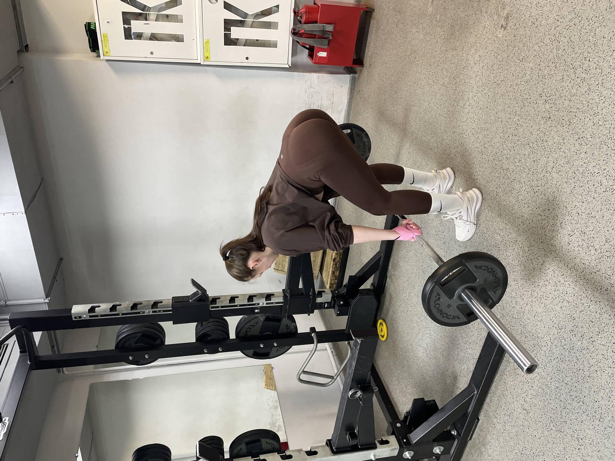 The image shows a woman in a gym in Izhevsk, Russia. She is bent over at the waist, holding a barbell. She is wearing brown leggings, a brown sweatshirt, white socks, and white sneakers. She is also wearing pink gloves. The barbell is loaded with weight plates. The gym has gray floors and white walls. There is a squat rack and weight storage to her left. There are white boxes affixed to the wall behind her.