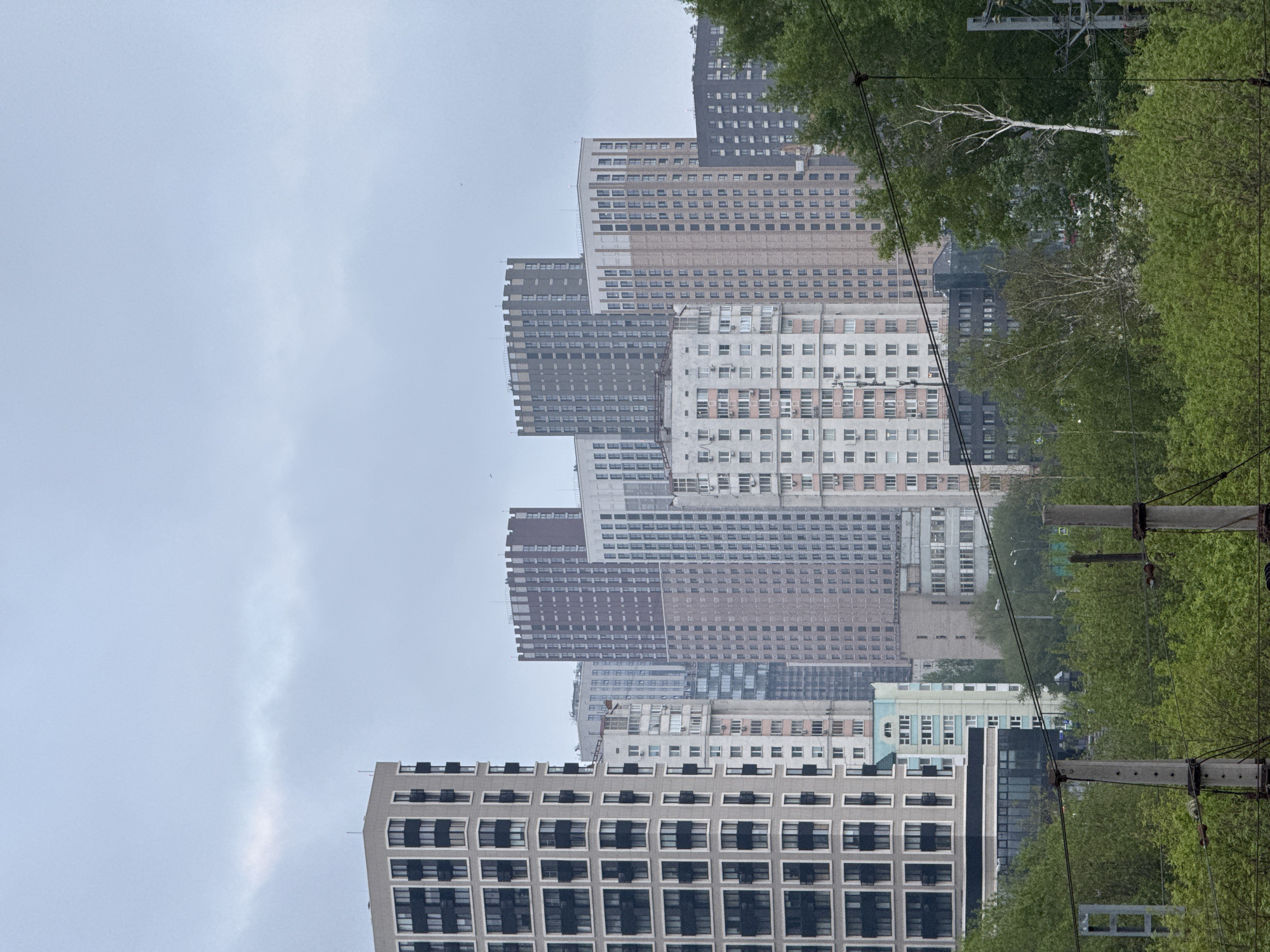 The image shows a view of multiple buildings in a cityscape, likely located in Moscow, Russia. The structures vary in height and design, ranging from older apartment blocks to newer high-rise buildings with modern facades. The weather appears overcast and misty, creating a hazy backdrop. Utility poles with electrical wires are visible on the right side of the image, partially obscured by trees with green foliage.
