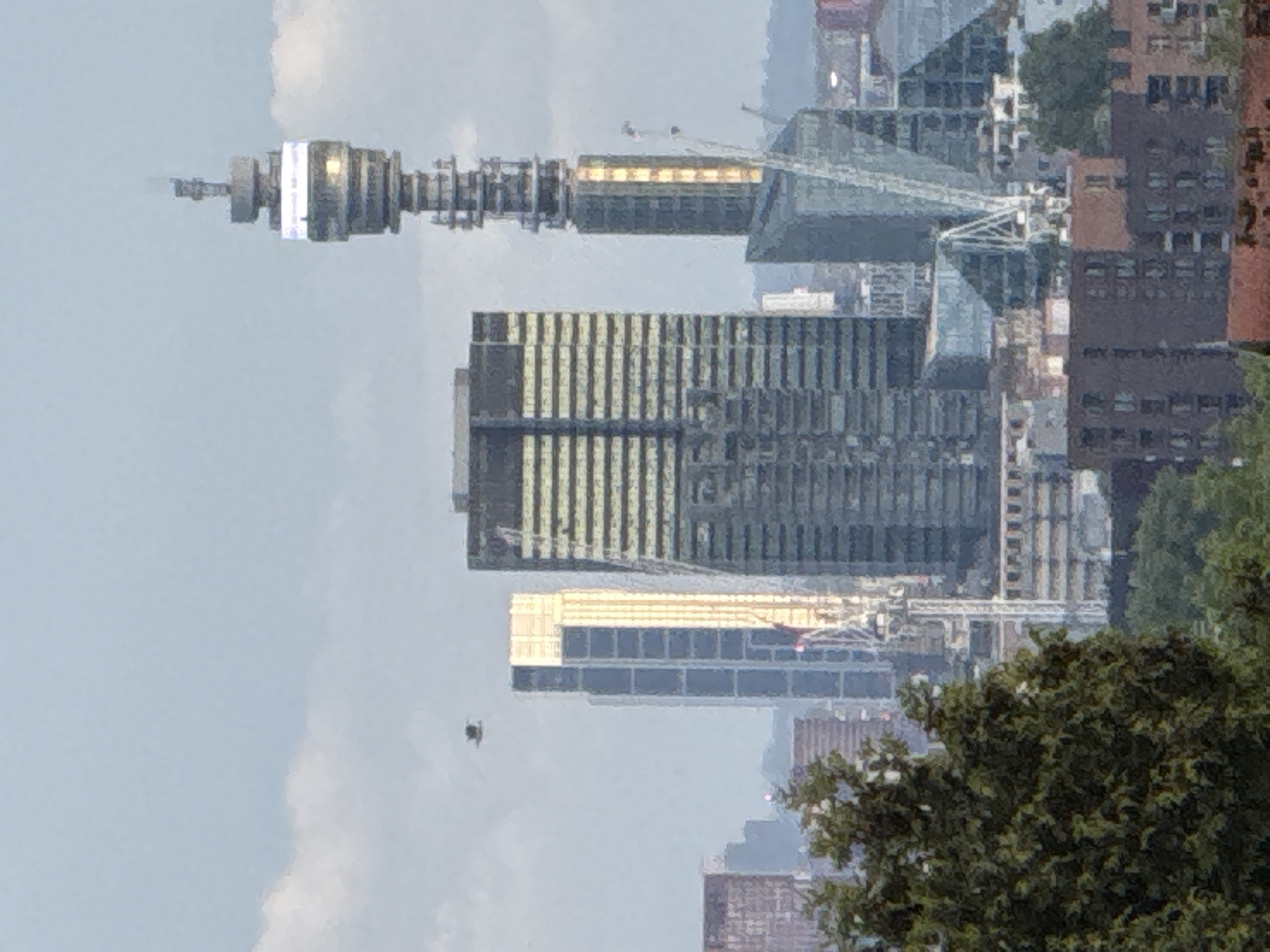 The image presents a view of the London skyline, featuring several prominent buildings.
In the left of the frame stands the BT Tower, recognizable by its distinctive cylindrical and tiered structure. Adjacent to it is a modern building with a complex angular design, likely a commercial or residential structure. Further to the right, a taller building with a grid-like facade is visible. A crane is also present, indicating ongoing construction in the area.
The right side of the image shows more dense urban development, including buildings of varying heights and designs, intermixed with green trees. The sky is overcast with some clouds.
Based on the architectural styles and the presence of the BT Tower, it can be deduced that the image captures a view of the city of London, United Kingdom, likely the area around Fitzrovia or Bloomsbury.