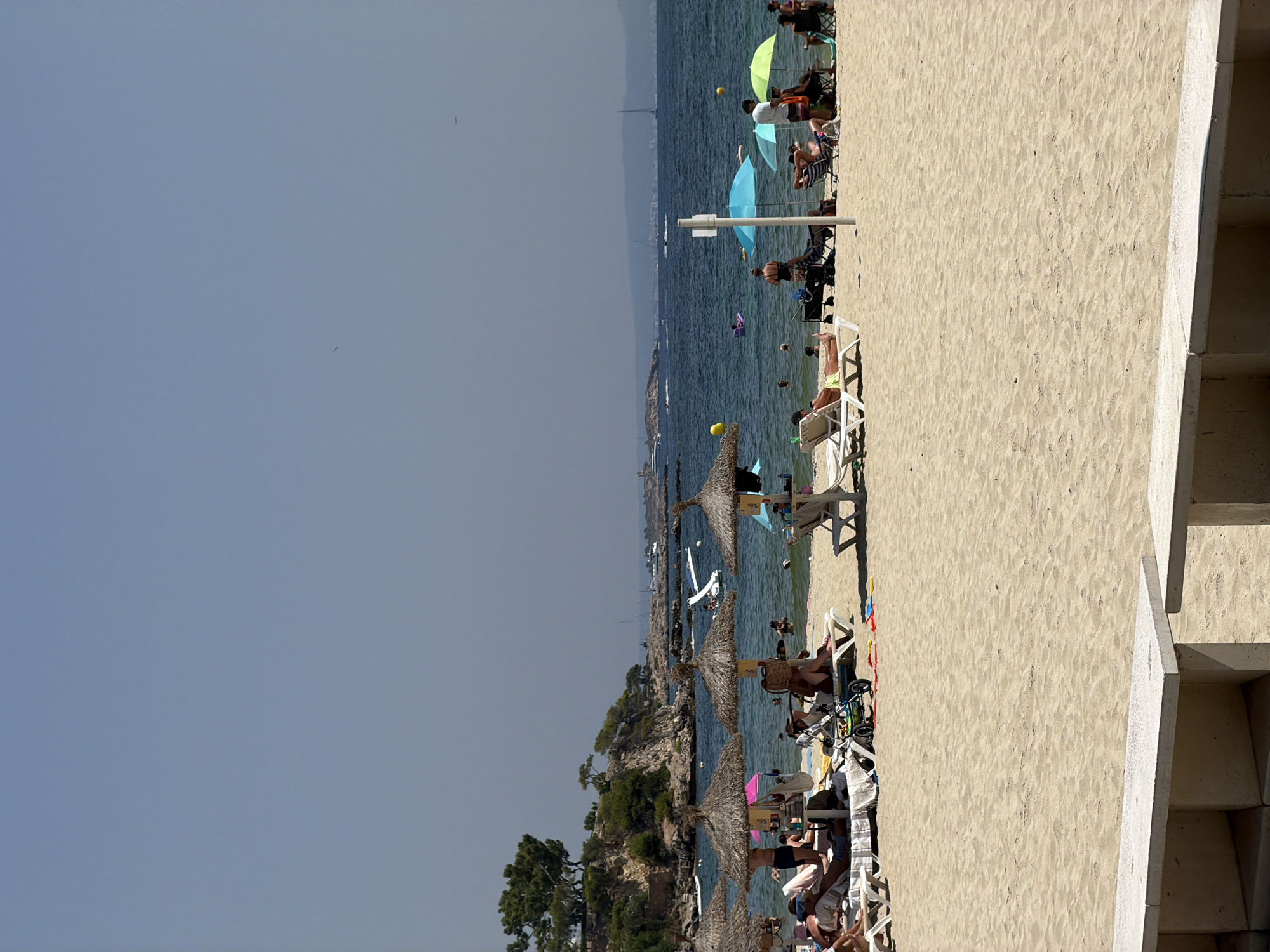 The image depicts a sunny day at Palmanova Beach, Spain. The foreground features a sandy beach with several beach chairs and umbrellas set up. In the mid-ground, people are sunbathing on loungers, some are in the shallow water, and others are walking along the shore. A baby stroller is visible near some of the chairs. The background shows the clear blue sea with a few boats and distant landmasses. The sky is clear and bright blue, indicating fair weather. The overall scene is a typical summer day at a popular tourist beach.