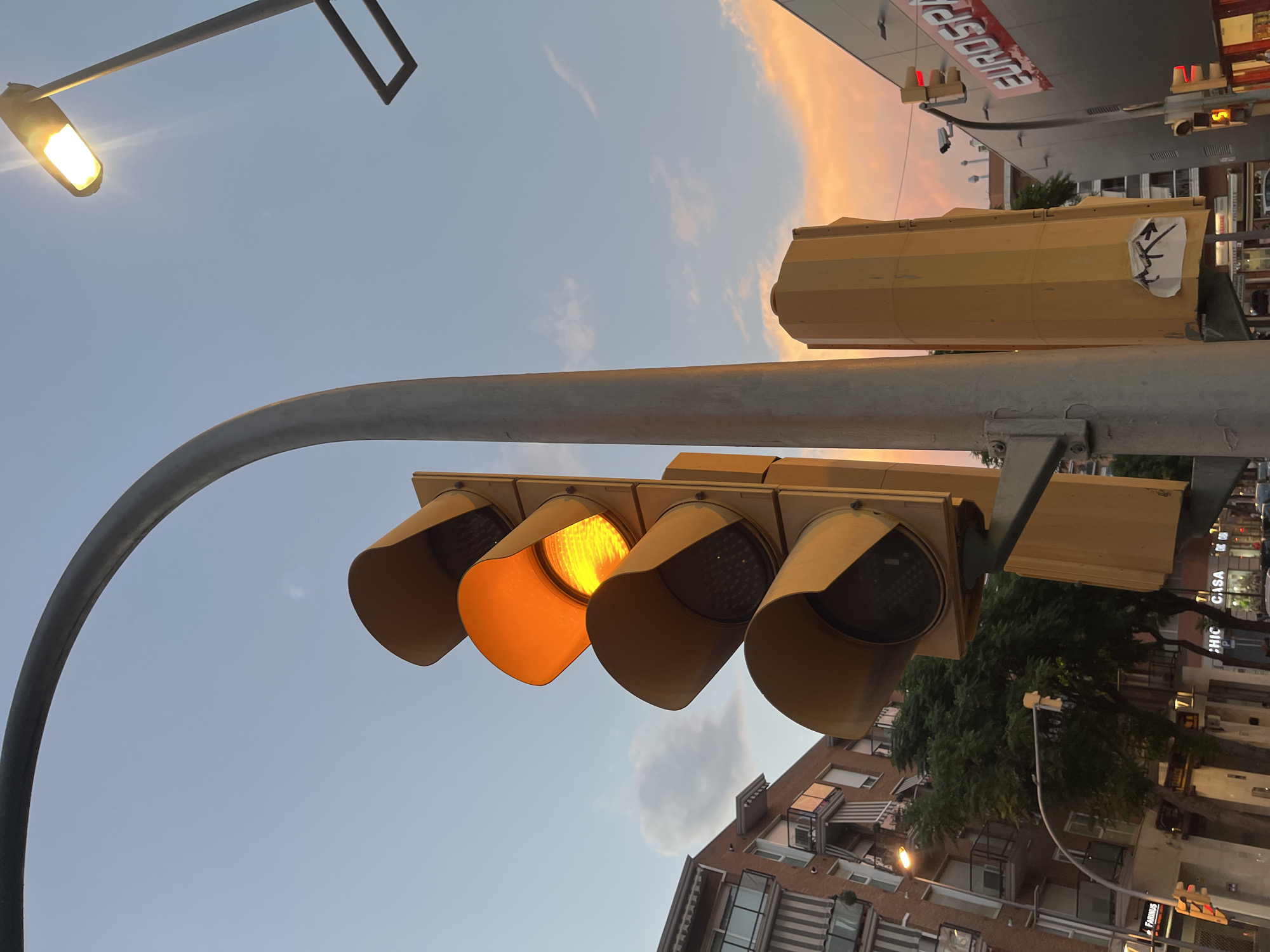 The image captures a traffic light illuminated with a yellow signal, positioned in the city of Tarragona, Spain. The traffic light is a prominent yellow structure with three lights stacked vertically; the middle light, indicating yellow, is lit brightly. Above and to the right of the traffic light, a street lamp with a bright light source is visible, casting a glow. The sky above is a clear blue with wispy clouds tinged with orange and pink, suggesting either dawn or dusk.
In the background, on the left side of the frame, a multi-story building with balconies and awnings is visible. Adjacent to the building, trees with lush green foliage can be seen. To the right of the traffic light, a modern building with the prominent red and white "EUROSPAR" signage is present. Further in the background, other buildings and street lights are discernible. The scene appears to be a street intersection, likely in an urban area, with typical city elements like buildings and street infrastructure. No people are clearly visible in the frame. The overall lighting suggests it's either early morning or late evening.