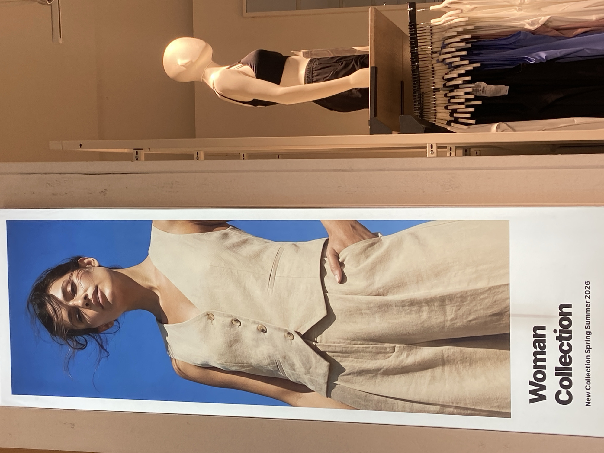 This image captures a retail display, likely within a clothing store in Budva, Montenegro. The photograph itself is rotated 90 degrees clockwise.
The upper section of the rotated image features a light-toned, headless mannequin positioned horizontally, dressed in a black sports bra and black shorts. Adjacent to the mannequin on the right, a clothing rack is visible, holding numerous garments on hangers, predominantly in shades of white, light blue, and black. The store interior around the mannequin and rack appears well-lit by warm artificial light.
The lower half of the image is dominated by a large, eye-catching poster or banner. It showcases a young woman with dark hair, depicted from the chest up, looking upward with a composed expression. She is wearing a light beige or linen-colored sleeveless top or vest with several prominent buttons down the front, paired with what appears to be matching wide-leg trousers. Her left hand is casually tucked into a pocket. The background behind her is a vivid, clear blue sky, indicating an outdoor, sunny environment with strong natural lighting casting shadows on her face.
Text is vertically aligned along the right edge of the poster, reading: "Woman Collection New Collection Spring Summer 2026." The image functions as an advertisement for this upcoming fashion collection.