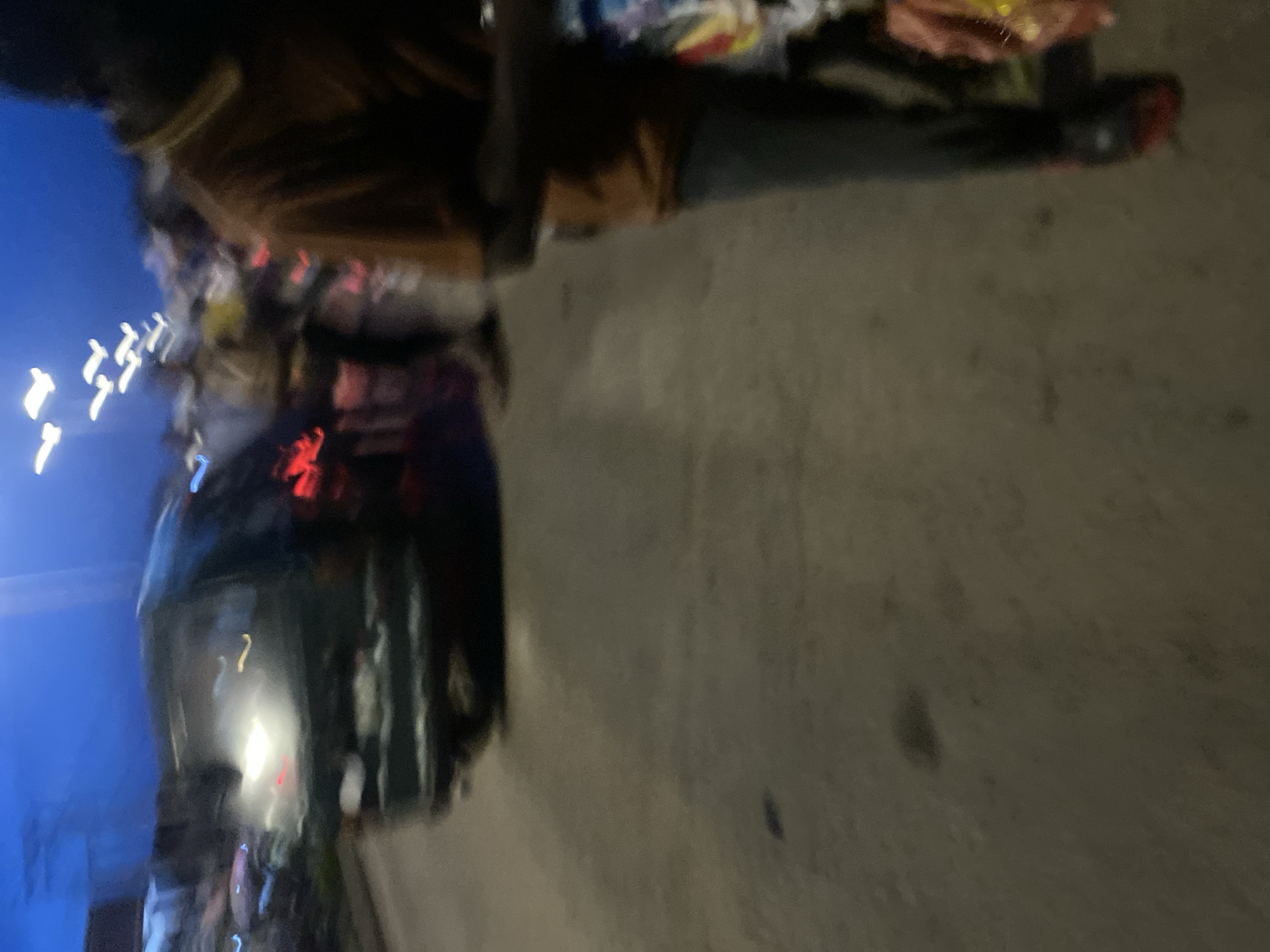 The image shows a street scene in Bukuru, Nigeria, at dusk. A dark-colored vehicle is visible in the background, with its headlights illuminated. Several figures, appearing to be pedestrians, are present on the street, some of whom appear to be vendors. The overall lighting suggests a low-light environment, likely due to the time of day. The image is blurry, indicating movement during capture.