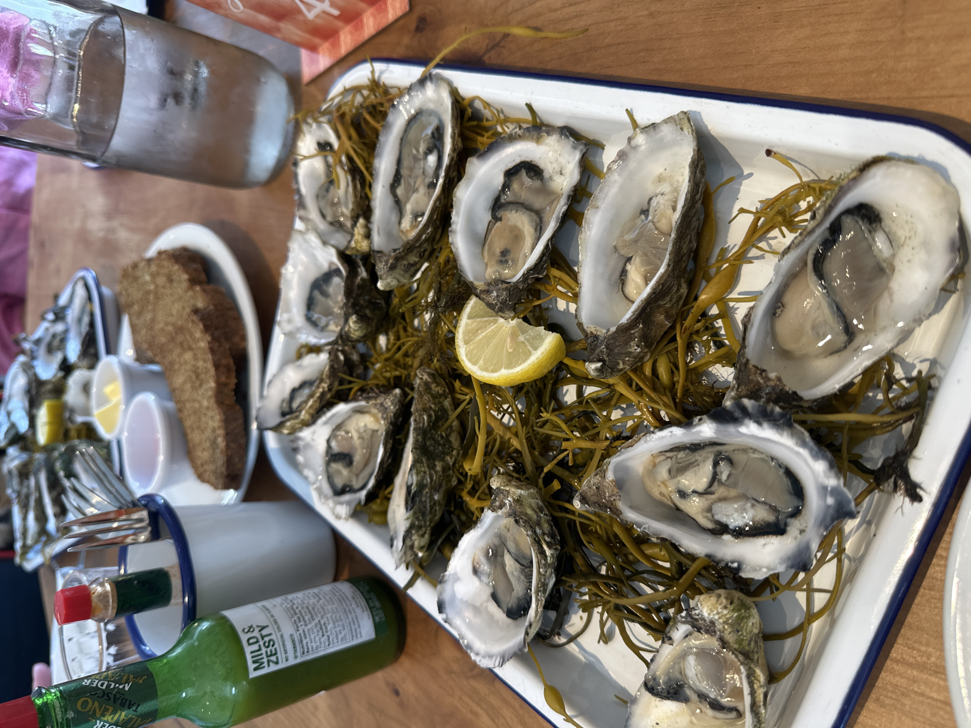 An overhead shot captures a table setting with a tray of fresh oysters, a bottle of Tabasco sauce, two small dipping sauce bowls, and bread. The oysters are served on a bed of what appears to be seaweed, arranged in a white enamel tray with a blue rim. A wedge of lemon rests on the tray near the oysters. In the background, a glass bottle filled with water and ice sits on a wooden table. The scene suggests a meal is being enjoyed at a restaurant in Rosduane, Ireland.