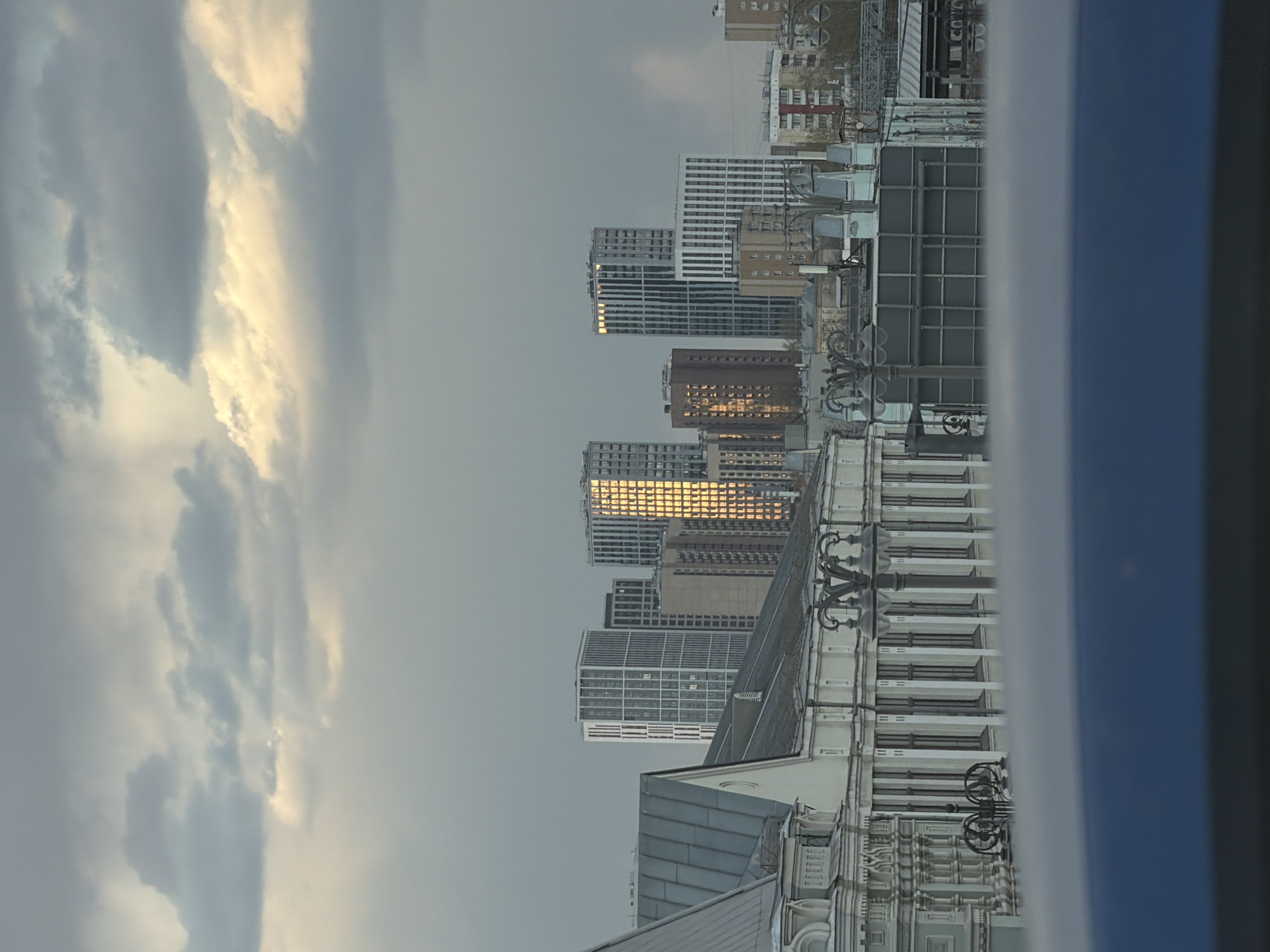 The image depicts a cityscape in Moscow, Russia, viewed from an elevated position, likely a window. The foreground shows a part of a building with ornate architectural details. The backdrop features a cluster of modern high-rise buildings reflecting the warm light of the sun, contrasting with the overcast sky filled with clouds. The buildings have a mix of white and gray facades, and some reflect the sun's glare. The overall environment suggests an urban setting with a mix of old and new architecture.