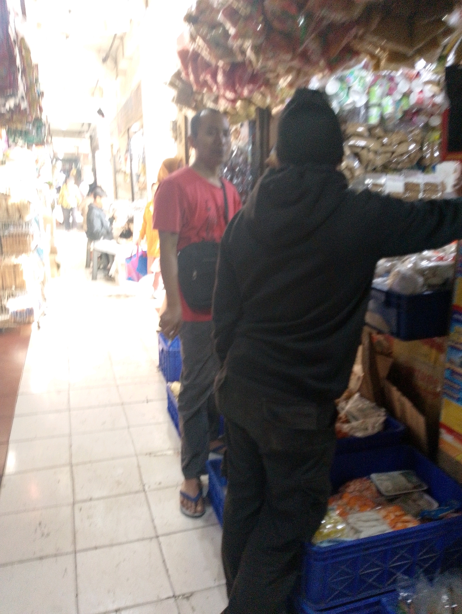 The image depicts an indoor marketplace scene, likely in Lembang, Indonesia, given the context provided. The foreground is dominated by two individuals. On the right, a person wearing a black hooded jacket and dark pants faces away from the camera, their back towards the viewer, obscuring their features. In the center, another individual wearing a red t-shirt and dark pants stands with their side partially visible. They are looking towards the left of the frame. The floor is tiled with off-white squares.
The background reveals a bustling marketplace with shelves and stalls lining a narrow aisle. Various goods are displayed, including packaged items and what appear to be decorative elements hanging from the ceiling. The lighting is bright and diffused, suggesting an open or well-lit interior. Several other blurry figures and shopping bags are visible further down the aisle, indicating a public space with shoppers and vendors. Blue plastic crates filled with merchandise are prominent in the lower right portion of the image. One crate contains a variety of packaged items, some appearing to be food-related with orange and white colors. Another crate to the left contains miscellaneous items. The overall impression is one of a lively and somewhat crowded market environment.