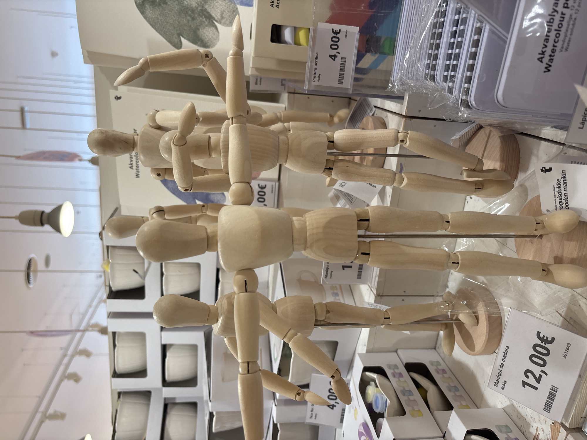 The image is a close-up shot of a retail display featuring several wooden artist mannequins. The mannequins are light-colored wood and are posed in various dynamic positions, with articulated limbs. They are positioned in the foreground and middle ground of the image.
In the background, shelves are stocked with white ceramic bowls and other products, suggesting this is likely a store that sells art supplies, home goods, or a combination of both. A sign partially visible in the background reads "Akvan Watercolor".
There are several price tags visible. One tag reads "Maniquí de madera Hobby 12,00€". Another tag reads "Pintura acrílica Hobby 4,00€". There is also a tag that says "Taza flor, D Hobby 4,00€".
The lighting appears to be from overhead fluorescent fixtures. The overall environment is a retail space.
While the image is taken in Orihuela, Spain, the exact location within the city is not discernable from the visual information provided. There are no people visible in the image. The focus is on the inanimate objects for sale.