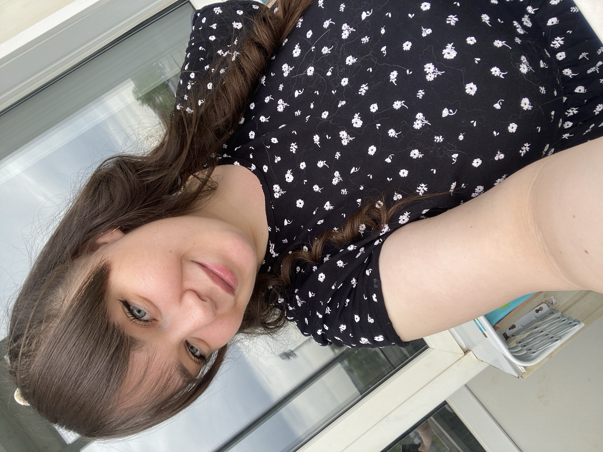 A woman with brown hair and blue eyes is taking a selfie indoors near a window. She is wearing a black dress with white floral patterns. The window frame is white, and reflections in the glass suggest an outdoor environment. No information indicates this photo was taken in Votkinsk, Russia.