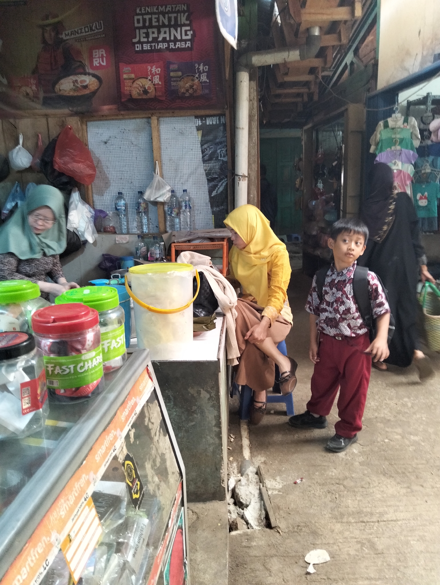 In Subah, Indonesia, a street-level outdoor market scene unfolds. A young boy, possibly around 8-10 years old, stands in the foreground, wearing a patterned short-sleeved shirt and maroon pants, with a black backpack on. To his left, a woman in a yellow hijab and long yellow top sits on a blue stool, her legs crossed, with a brown skirt. Behind her, another woman in a green hijab is partially visible. Various market stalls are present, displaying goods such as packaged instant noodles with Japanese text, bottled water, and jars of food items with prominent "FAST CHAR" labeling. A glass display counter with various items, possibly phone accessories, is also visible. The ground is a mixture of concrete and soil, with some debris and a small hole. The overall impression is one of a bustling, everyday market environment.