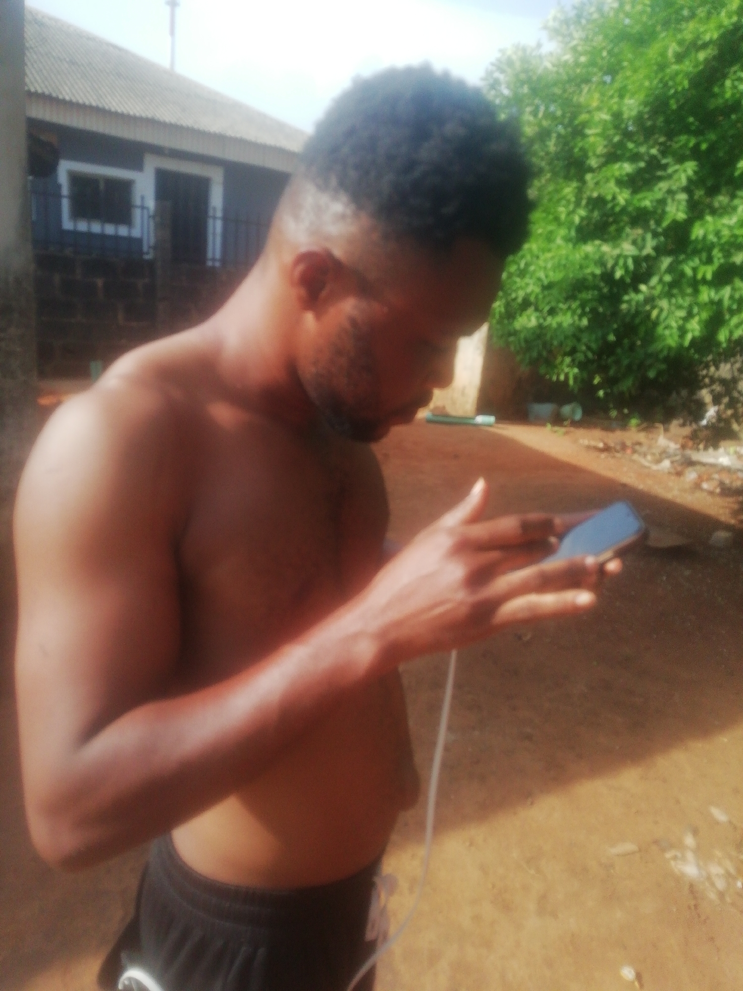 The image depicts a shirtless man standing outdoors, looking at a mobile phone he is holding. He has dark hair, a beard, and is wearing shorts. A charging cable is attached to the phone. Behind him, there's a building with a corrugated iron roof and a small window, and a lush green tree or bush is visible. The ground appears to be dirt or gravel. The location is in the city of Unknown, Unknown.