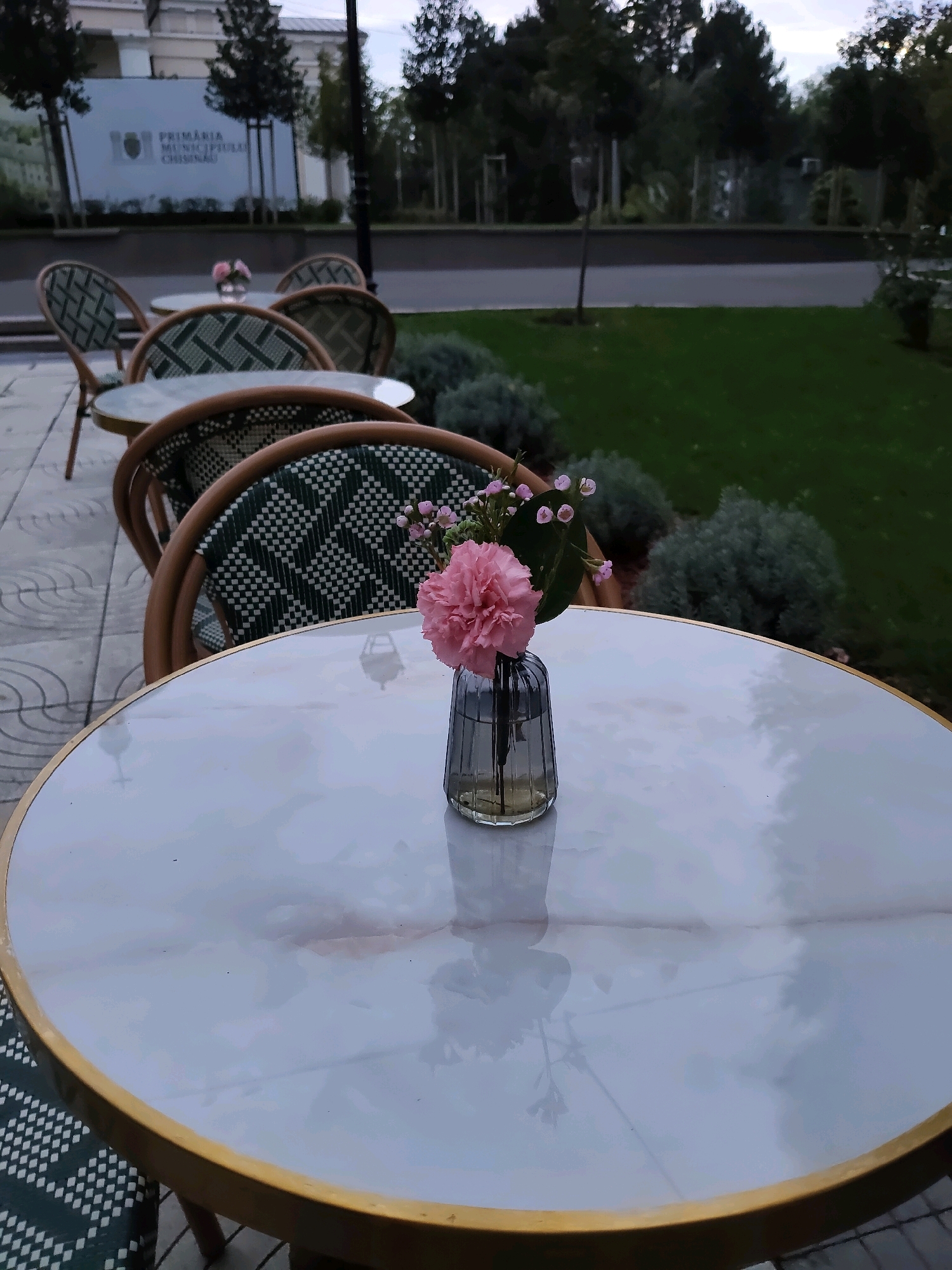 The image shows an outdoor seating area with several tables and chairs. A single table in the foreground has a small vase with a pink flower and some smaller pink blossoms. The chairs have a distinctive green and white checkered pattern. In the background, a sign can be seen which reads "PRIMĂRIA MUNICIPIULUI CHIȘINĂU," indicating the location is in Chișinău, Moldova. The setting appears to be a cafe or restaurant patio adjacent to a grassy area with trees. The sky is overcast, suggesting it might be evening or a cloudy day. No people are visible in the image.