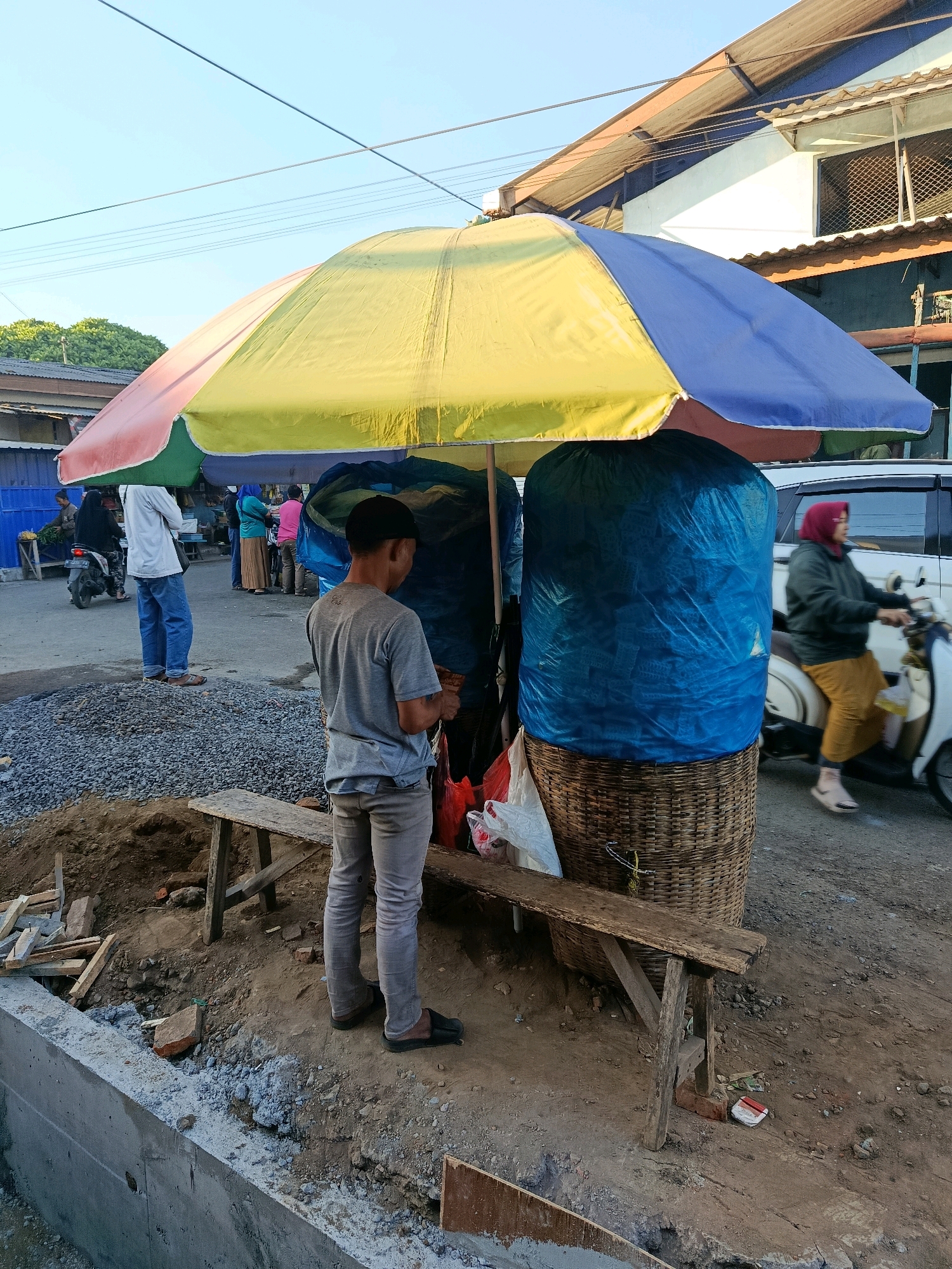 A young boy wearing a grey t-shirt and grey pants stands under a large, multi-colored umbrella at a street stall in Bululawang, Indonesia. The umbrella is predominantly yellow, blue, red, and green. The stall features a large, blue tarp-covered object, possibly goods for sale, placed on top of a woven basket. A rough wooden bench is situated in front of the stall. In the background, other people are visible, including a man in a white shirt and blue jeans, and a woman riding a scooter wearing a red headscarf and yellow pants. The environment appears to be a street with some construction materials, including gravel and concrete. The time of day seems to be late afternoon, with the sun casting long shadows.