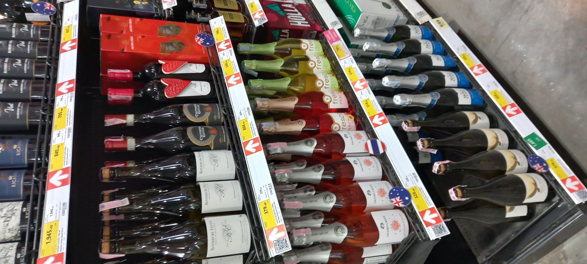 This image captures a well-stocked aisle within a supermarket or liquor store, likely located in Moo 1, Thailand. The scene displays multiple shelves filled with various bottles of wine and sparkling wine, neatly arranged in vertical and horizontal rows.
The products include a range of wines such as red wines (e.g., "CABERNET S" from "RICASON" and "ROBERT MO"), white wines ("ROTHBURY ESTATE"), rosé, and sparkling wines, some of which appear to be fruit-flavored like "fresh PEACH." Prominently featured brands include "RICASON," "ROBERT MONDAVI," "ROTHBURY ESTATE," and "JACOBS CREEK."
White price tags with red directional arrows are affixed to the shelves, indicating prices and product locations. These tags display prices such as 1,945.00, 845.00, 705, 687, 804, 379.00, 798, 540, 214, 528, 378, and 1,485, which, given the location, are likely in Thai Baht. Some price tags also feature QR codes. Small circular flags are visible on some bottles, with an Australian flag seen on a Jacob's Creek bottle and a blue, white, and red vertical-striped flag, possibly French, on a sparkling rosé bottle. The overall setting is an indoor retail environment, designed for product display and sale. No people or specific activities other than product merchandising are depicted.
