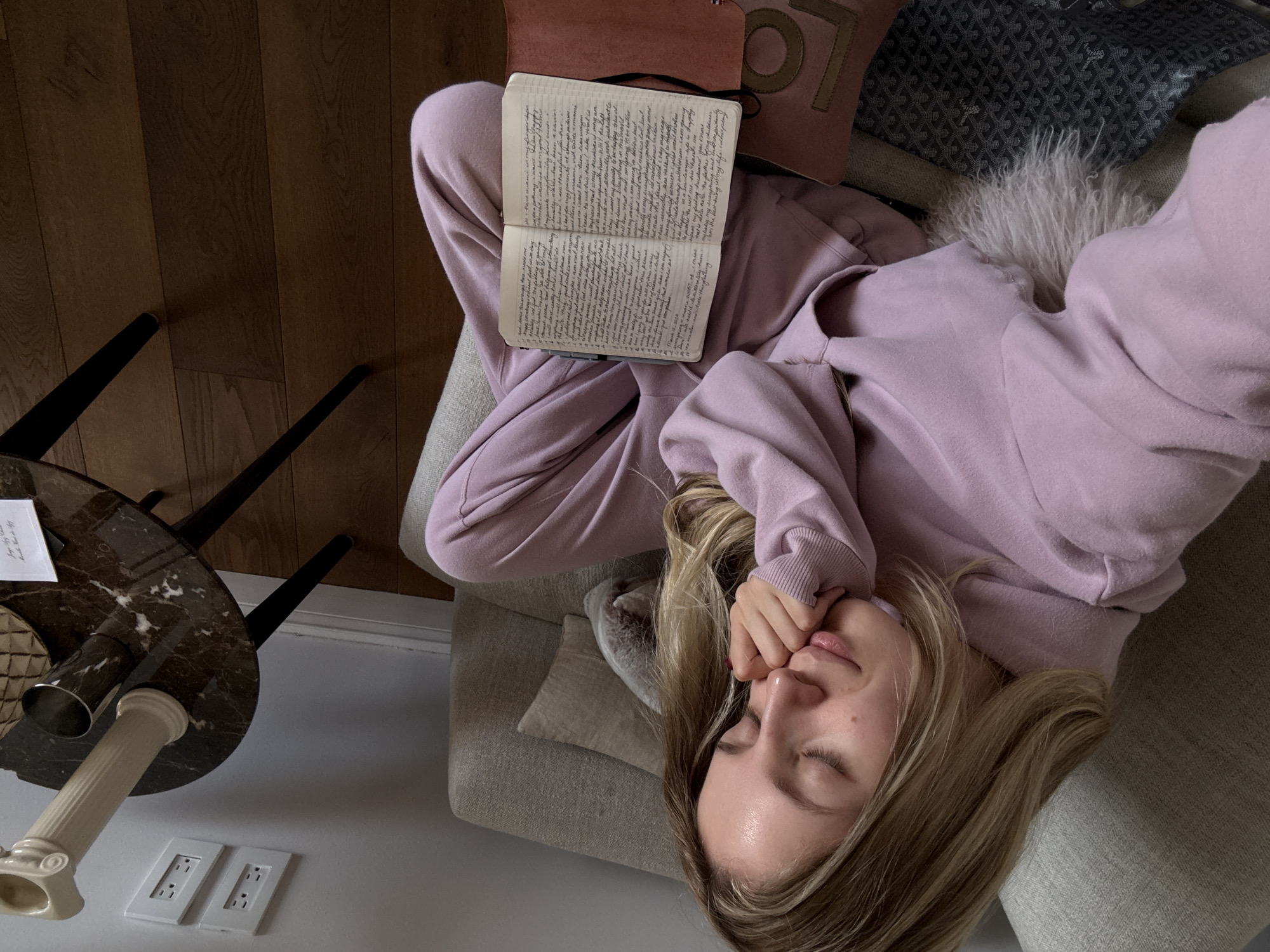 The image depicts an indoor, residential scene in Mexico City, Mexico. A blonde female is seated on a light grey fabric couch, leaning back with her eyes closed, suggesting rest or sleep. She wears a lavender-colored hooded tracksuit. Her right hand is positioned near her mouth, with fingernails visible. An open notebook filled with handwritten text and a dark pen is resting on her lap, covering part of a dark brown object with the letters "LO" visible, possibly a bag or journal cover. A grey patterned object is partially visible beneath her left leg. To the right of the woman, a round dark brown marble-top side table with black legs holds a cream-colored decorative column, a silver metal cup, and a small white piece of paper. The background wall is light grey, featuring two white rectangular electrical outlets mounted vertically. The floor consists of light brown hardwood planks.
