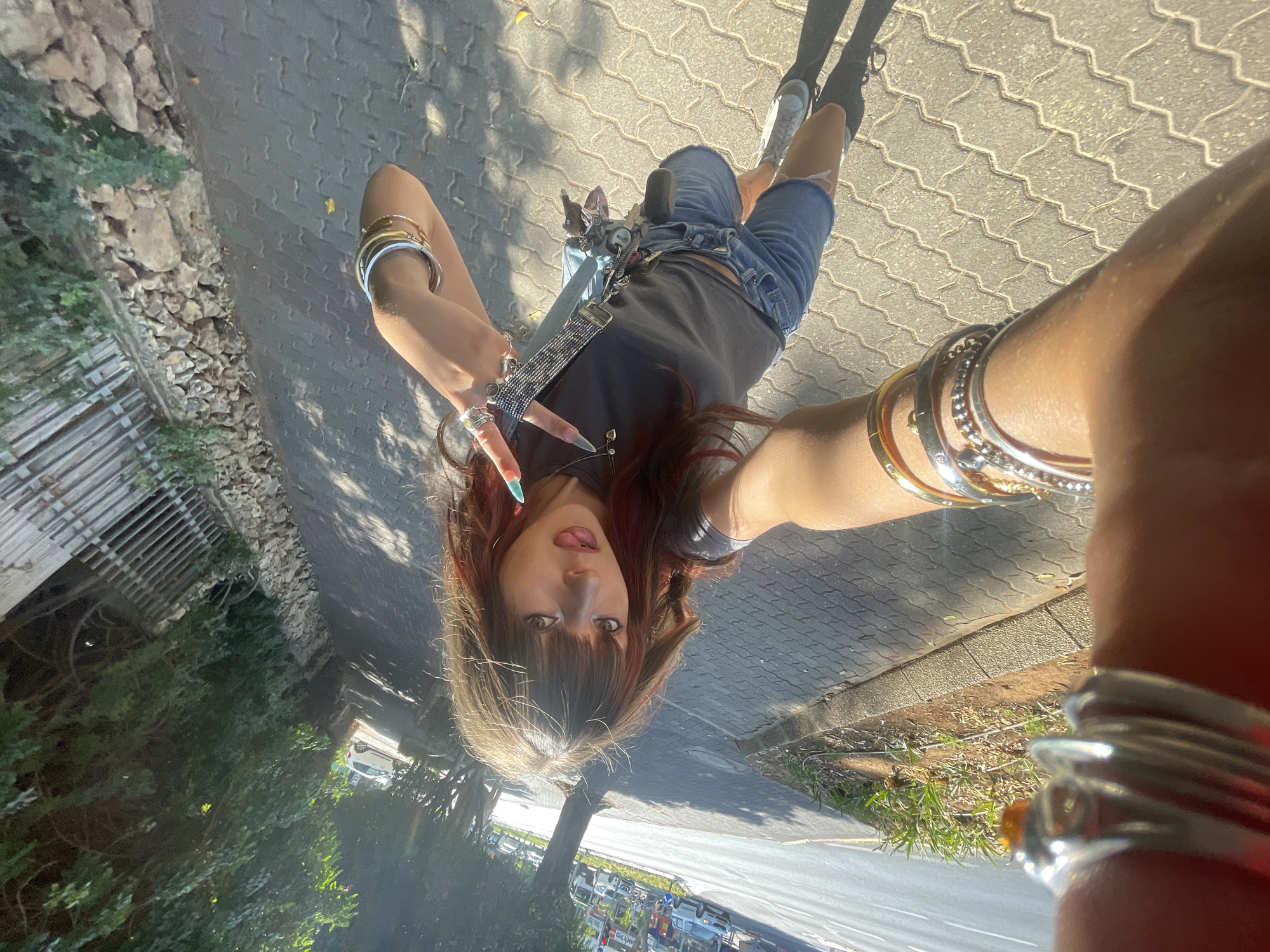 A young woman with long, auburn-highlighted hair and bangs stands on a sidewalk, holding a phone up to take a selfie. She is wearing a dark gray t-shirt, denim shorts, and white sneakers. She has several bracelets on her wrists and rings on her fingers. Her tongue is sticking out, and she is making a peace sign with her left hand. The background shows a paved sidewalk, a street with traffic, trees, and a stone wall. The lighting suggests it is a sunny day. The image appears to be taken in Raanana, Israel, given the style of the sidewalk paving and the presence of typical Israeli street foliage and architecture.