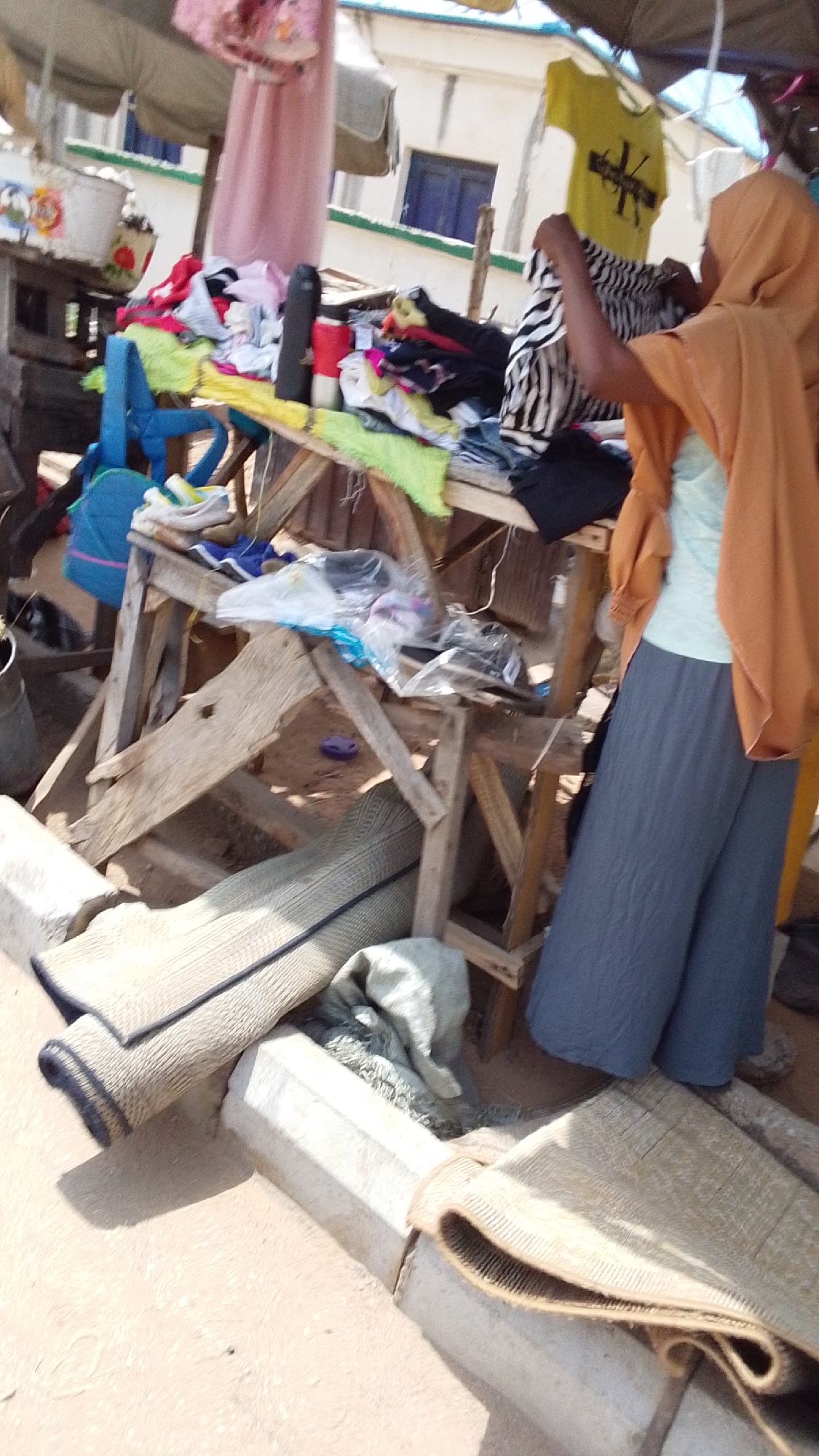 The image depicts an outdoor market stall in Lokoja, Nigeria. A woman wearing a hijab and an orange shawl is examining a zebra-striped garment on a wooden table laden with various clothing items. Behind the table, there is a collection of other items, including bags and bottles. The stall itself is a makeshift structure with wooden supports. In the foreground, there are woven mats rolled up next to a concrete curb. The overall impression is one of a bustling, informal marketplace.