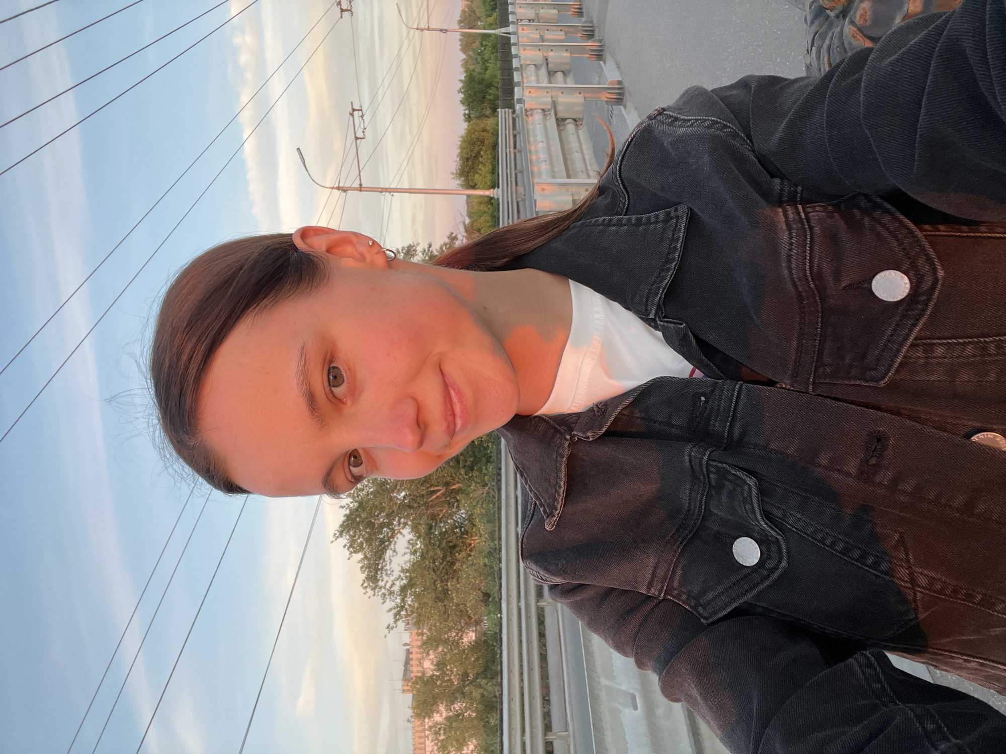 The image is a selfie taken by a young woman outdoors, likely on a bridge given the railing and road visible in the background. She has brown hair pulled back and is wearing a dark denim jacket over a white t-shirt. The setting appears to be urban, with buildings, trees, power lines, and streetlights visible in the distance. The lighting suggests it is either sunrise or sunset, with warm golden light illuminating the woman's face. The sky is a pale blue with some wispy clouds. The background shows a cityscape with various structures and vegetation. The image quality is clear, with no apparent digital manipulation. The location is identified as Novokuybyshevsk, Russia.