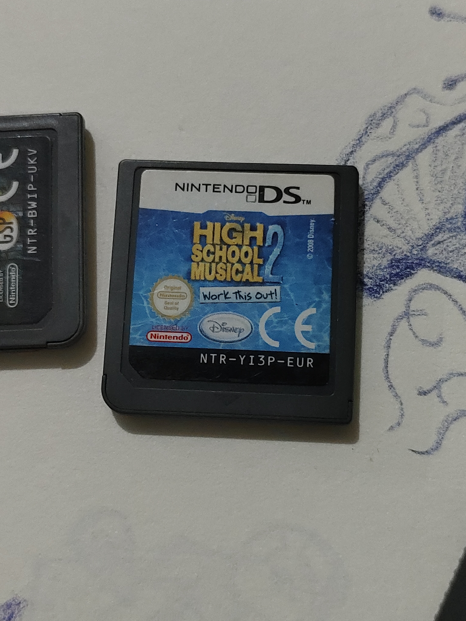 Two Nintendo DS game cartridges are positioned on a light-colored, textured surface adorned with blue scribbles, in the city of Unknown, Unknown. The cartridge on the left is partially visible, displaying the text "GSP", "NTR-BWIP-UKV", and "LICENSED BY Nintendo" on its side label. The prominent cartridge on the right features a full label for the game "HIGH SCHOOL MUSICAL 2: Work This Out!", bearing the "NINTENDODS™" logo at the top. Other visible elements on this label include a small Disney logo, "© 2008 Disney.", the "Original Nintendo Seal of Quality", "LICENSED BY Nintendo", a "CE" mark, and the cartridge code "NTR-YI3P-EUR". Both cartridges are dark gray or black plastic. The background surface is consistent in texture and tone, with abstract blue line drawings occupying the right and lower portions of the frame. No individuals are present, and no discernible actions or interactions are occurring.