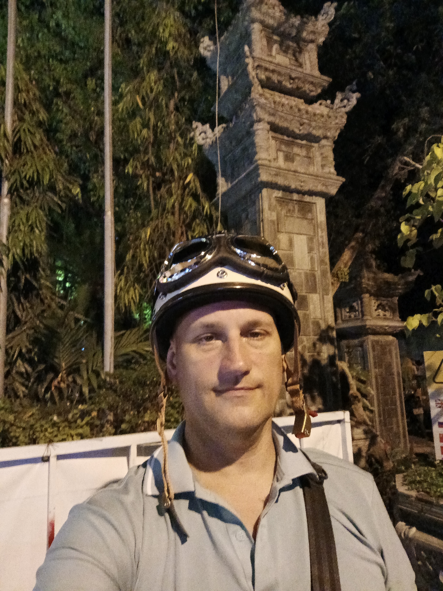 A man wearing a helmet with goggles stands in Tay Nha Trang Ward, Vietnam. He is facing forward, taking a selfie. Behind him is a stone structure, possibly an entrance to a temple or historic site. Vegetation and trees surround the scene, illuminated by artificial light sources. A white fence or barrier is also visible. The lighting is dim, suggesting it is nighttime. The man is wearing a light-colored polo shirt with a dark strap across his body.