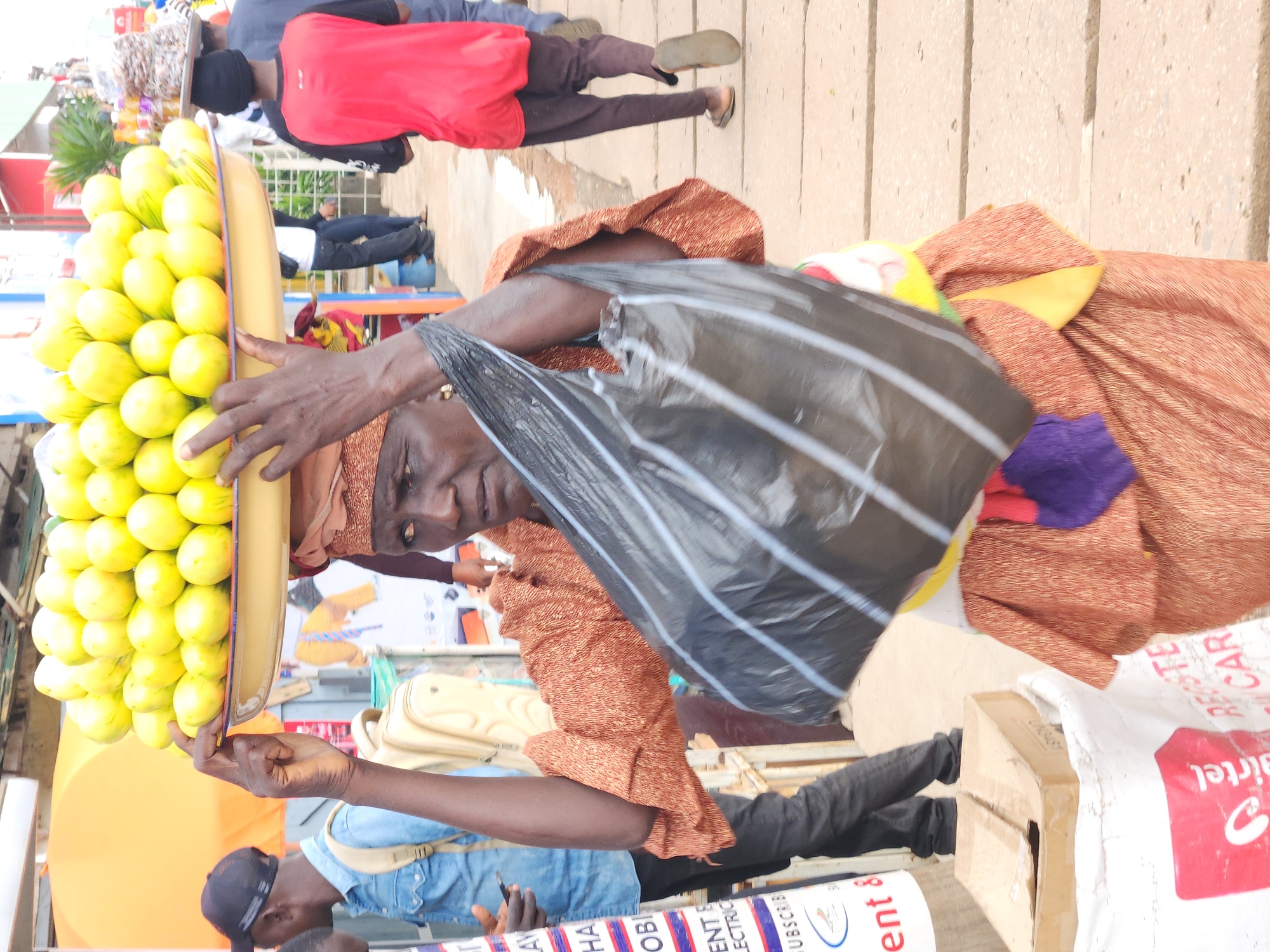 An elderly African woman is depicted in a medium shot, facing forward and slightly to her left, carrying a large yellow platter of bright yellow-green fruits on her head. She is dressed in a patterned orange fabric wrapped around her torso and lower body, with a dark head covering. Her right arm is raised, supporting the platter, and her left hand is holding a large black plastic bag. Her face shows a determined expression, with dark skin and visible wrinkles. The background is blurred but suggests an outdoor market setting in a city, with other people and stalls visible. The ground appears to be paved with concrete. The overall impression is of a vendor at work in a bustling environment.