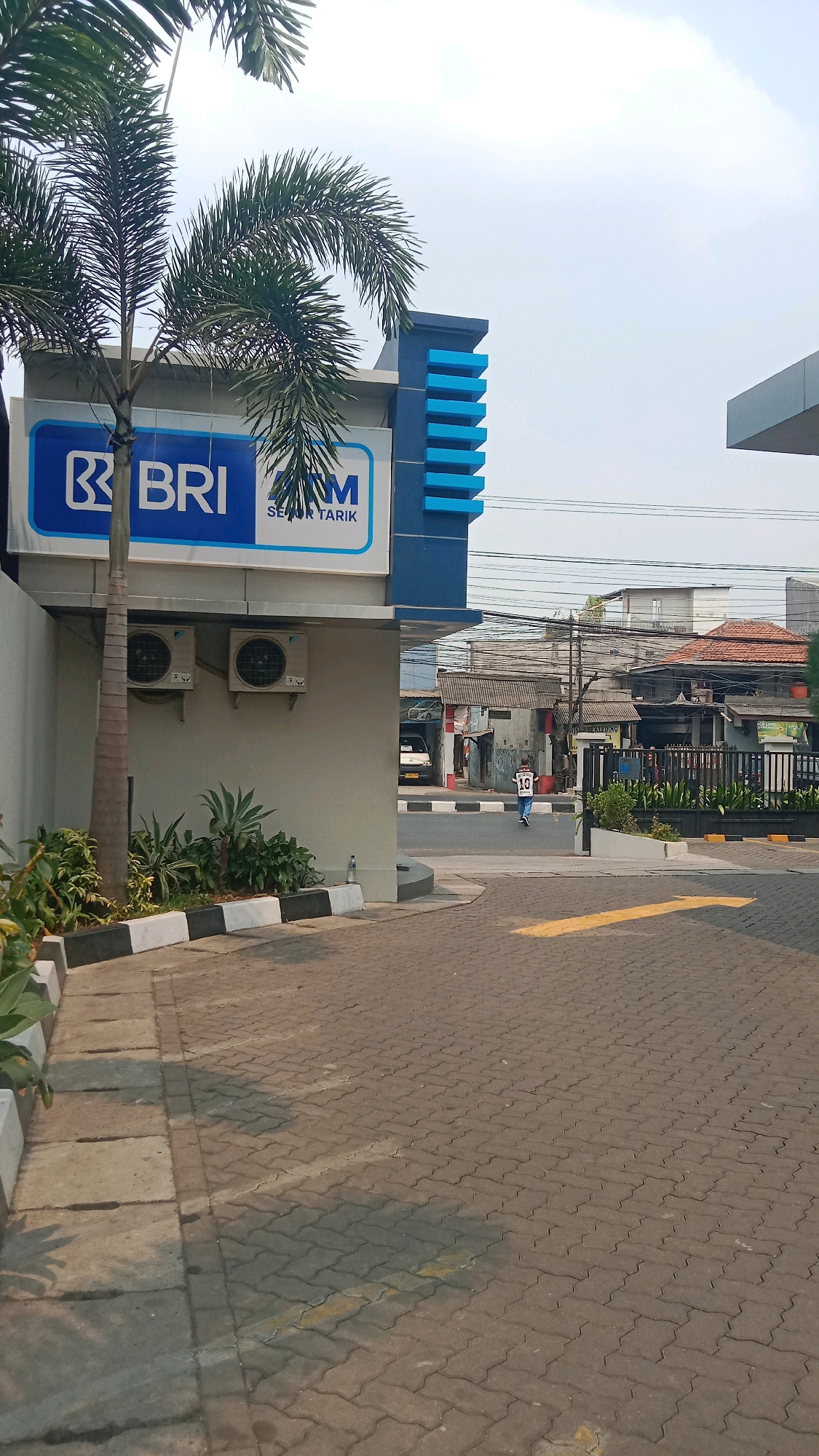 An outdoor shot features a building with a blue and white facade, displaying the logo "BRI" and "ATM". Two air conditioning units are mounted on the exterior wall. A palm tree is positioned in the foreground to the left. The ground is paved with interlocking bricks, and a yellow arrow is painted on it, indicating direction. In the background, there are several buildings, some with tiled roofs, and electrical wires crisscross the sky. A person wearing a white shirt with the number "10" on the back is walking away from the camera. The scene appears to be in a commercial or residential area in Bekasi, Indonesia, given the architecture and signage.