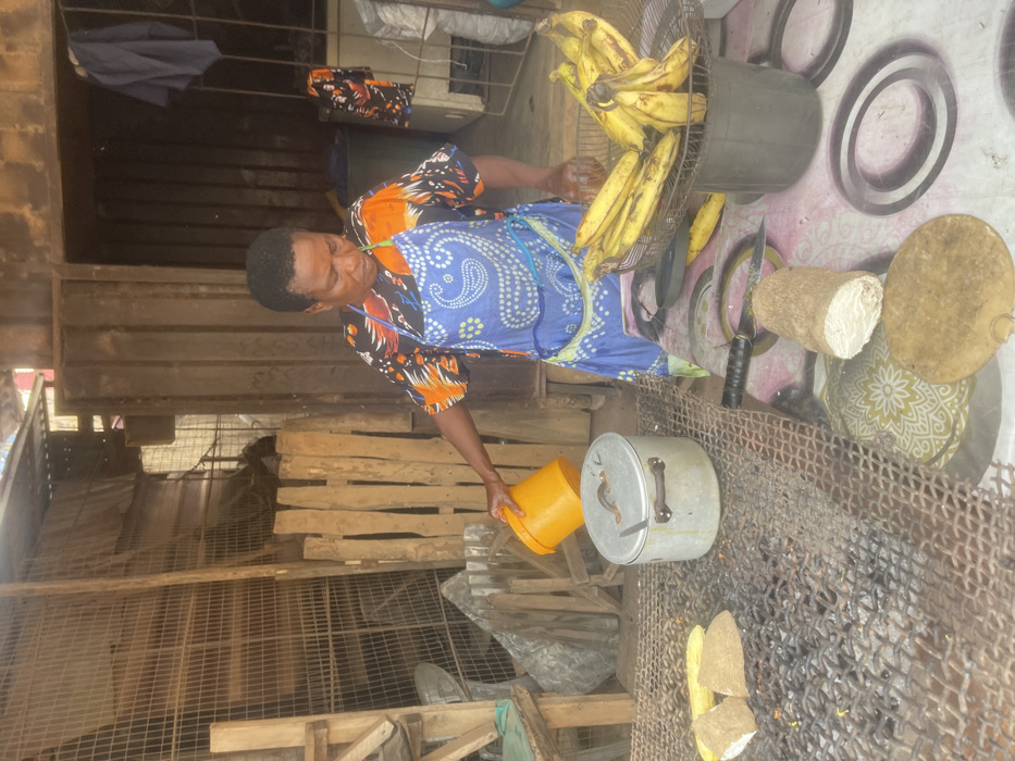 The image captures a scene featuring a woman engaged in food preparation, likely outdoors in a market or roadside stall setting. The image is oriented vertically, so describing it as if it were rotated 90 degrees clockwise would present a more natural viewing perspective.
In the center, a woman with dark skin and short, dark hair is seen from the chest up, looking downwards towards an aluminum pot. She is wearing a patterned short-sleeved top with orange, black, and white designs, and a blue apron with a white paisley-like pattern tied around her waist. Her right arm is extended, holding a bright yellow plastic bucket, from which she appears to be pouring liquid or contents into the open aluminum pot below. The pot has a lid with a handle resting beside it on the mesh surface.
To the right of the woman and slightly behind her, a large bundle of ripe yellow bananas is piled within what looks like the cage or guard of a standing fan, perhaps repurposed as a container. Further to the right, on a surface covered with a patterned mat (light pink with dark ring-like patterns), are several items. These include a large knife with a black handle, a cylindrical object made of a light fibrous material (possibly a type of root vegetable or processed food like fufu dough), and a round, flat wooden cutting board. Another patterned fabric or mat with a green and white design is also visible underneath some of these items.
In the foreground, spanning the lower part of the image, is a robust metal mesh or grill, possibly used for cooking over an open fire, as some dark, burnt residue and small glowing embers are visible beneath it, suggesting a charcoal grill. On this grill, in front of the pot, are pieces of what appear to be plantain or cassava, split lengthwise and ready for cooking or roasting.
The background consists of various wooden structures, including planks and what looks like a rudimentary wooden fence or stall framework. Parts of the ground are visible, appearing somewhat uneven and dusty. Overlapping with the top left of the image, some dark blue and patterned fabrics are hanging on a line or rail. The lighting suggests daytime, possibly bright sunlight, given the visible shadows and overall illumination. No discernible text is visible in the image.