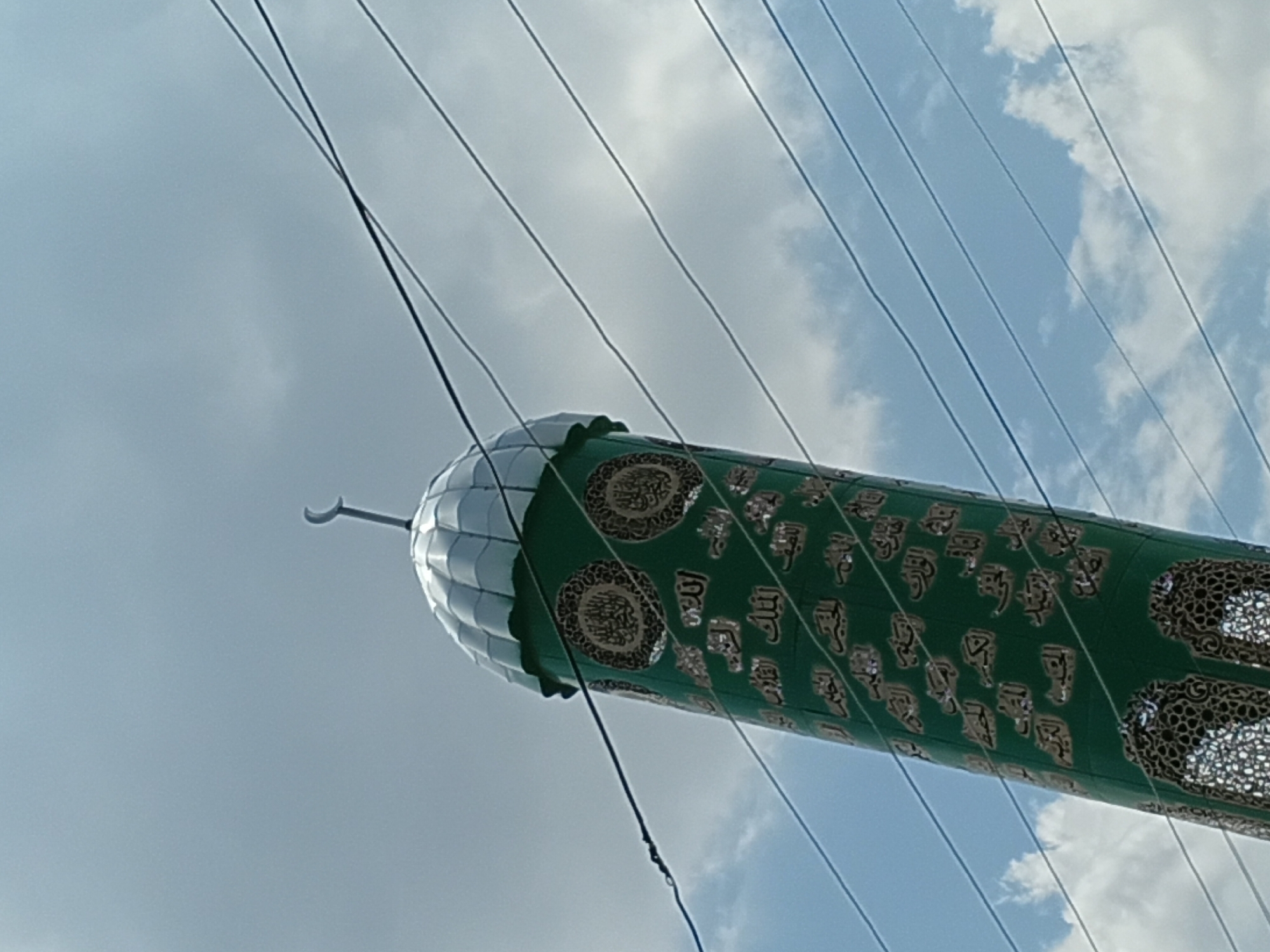 The image shows a close-up, low-angle shot of a minaret in the city of Osh, Kyrgyzstan. The minaret is cylindrical and painted green, adorned with intricate gold Arabic calligraphy and decorative patterns. It is topped with a silver, dome-like structure with a crescent moon finial. Several dark power lines crisscross the frame in front of the minaret and the sky. The sky is partly cloudy with patches of blue visible. The minaret appears to be part of a mosque, a prominent religious structure in Osh. The calligraphy on the minaret is likely verses from the Quran or the names of Allah.
