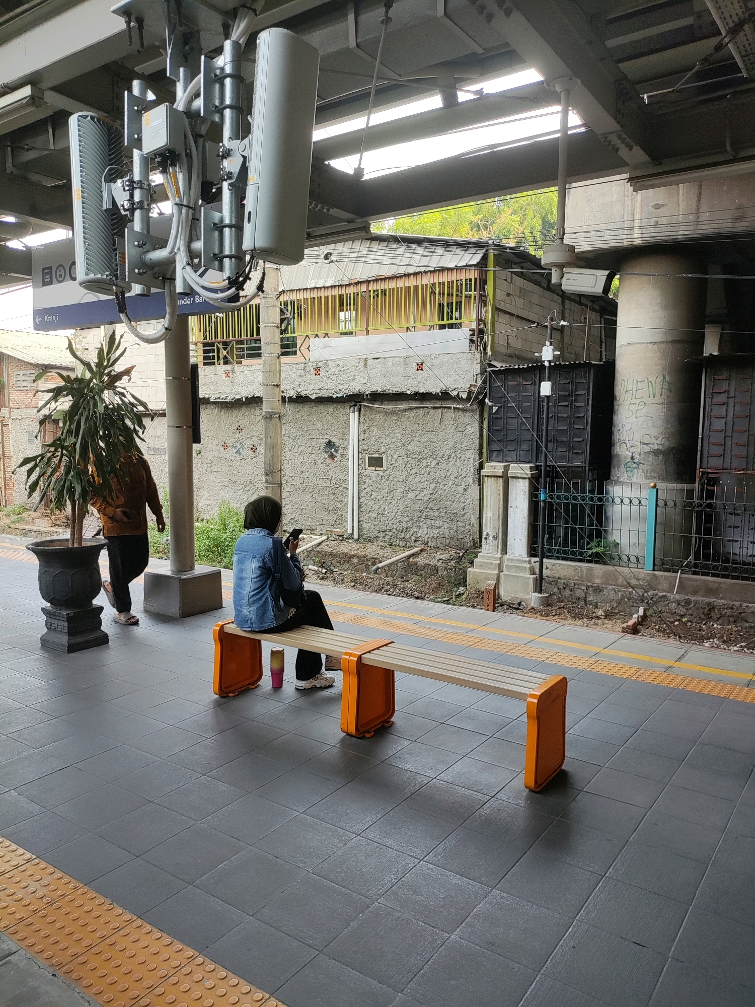The image depicts a railway station platform in Bekasi, Indonesia. In the foreground, a woman wearing a denim jacket and hijab is seated on an orange and beige bench, looking at her smartphone. To her left, a person wearing a brown jacket and dark pants walks away from the camera. A large potted plant stands near the person walking away. Overhead, a complex network of communication equipment, possibly for cellular service, is mounted on the station's structure. Further in the background, a security camera is visible mounted on a concrete pillar. Buildings and fencing line the area behind the platform. The platform itself is paved with dark gray tiles, with a yellow tactile paving strip along the edge.