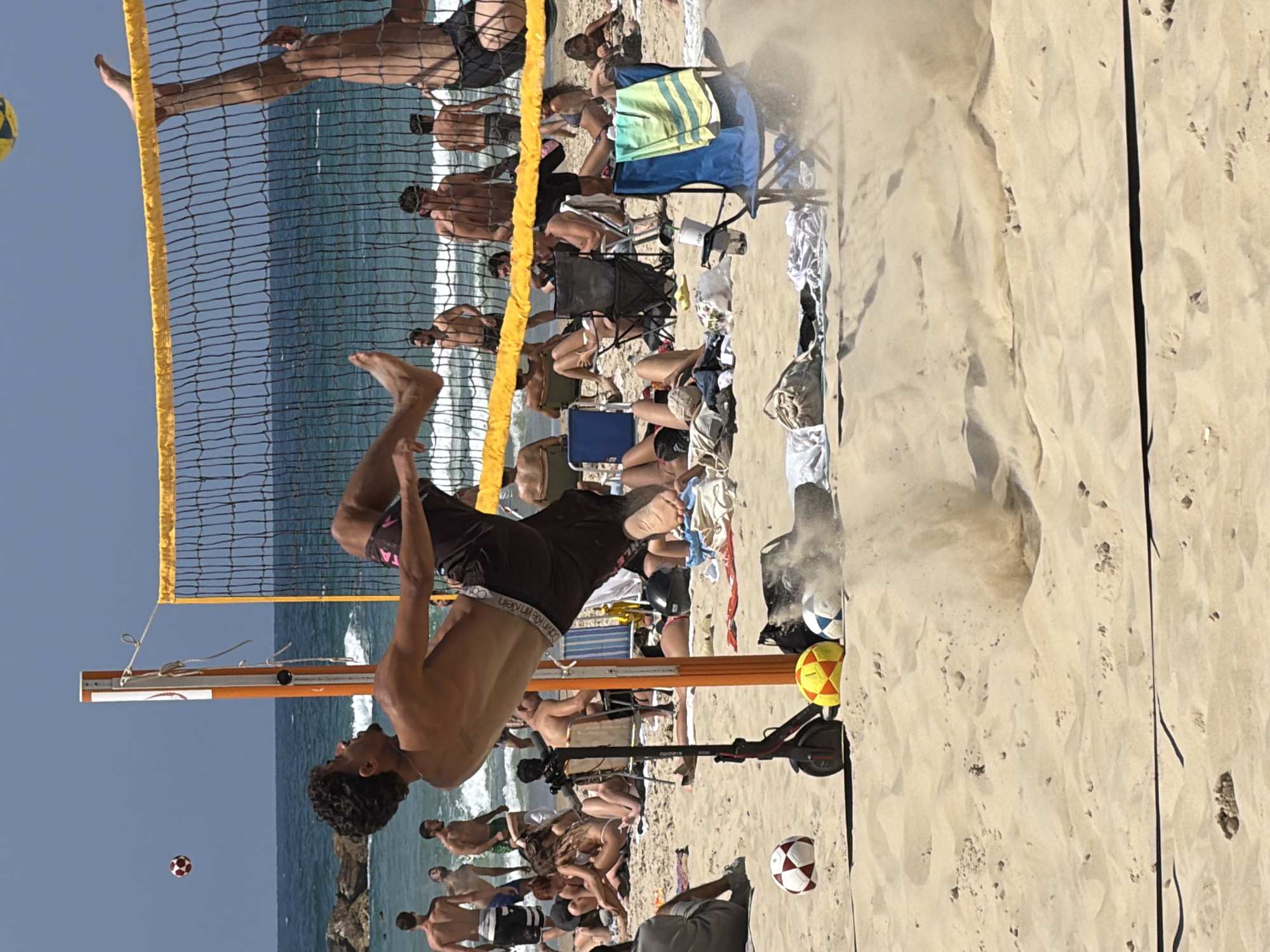 The photograph captures a vibrant beach scene, likely in Tel Aviv, Israel, due to the ocean view, the architectural elements of the buildings in the distance, the coastal geography, and the style of dress and activity of the people.
A beach volleyball net spans the scene, partially obscuring the ocean. A man is in mid-air, executing a backflip, presumably playing volleyball. Several other people are visible, some seated in beach chairs or on the sand, and others standing or swimming in the ocean.
Near the man executing the backflip, there is a black scooter. There are two soccer balls on the beach, one near the scooter.
The weather appears sunny, and the overall scene suggests a lively beach environment in Tel Aviv.
