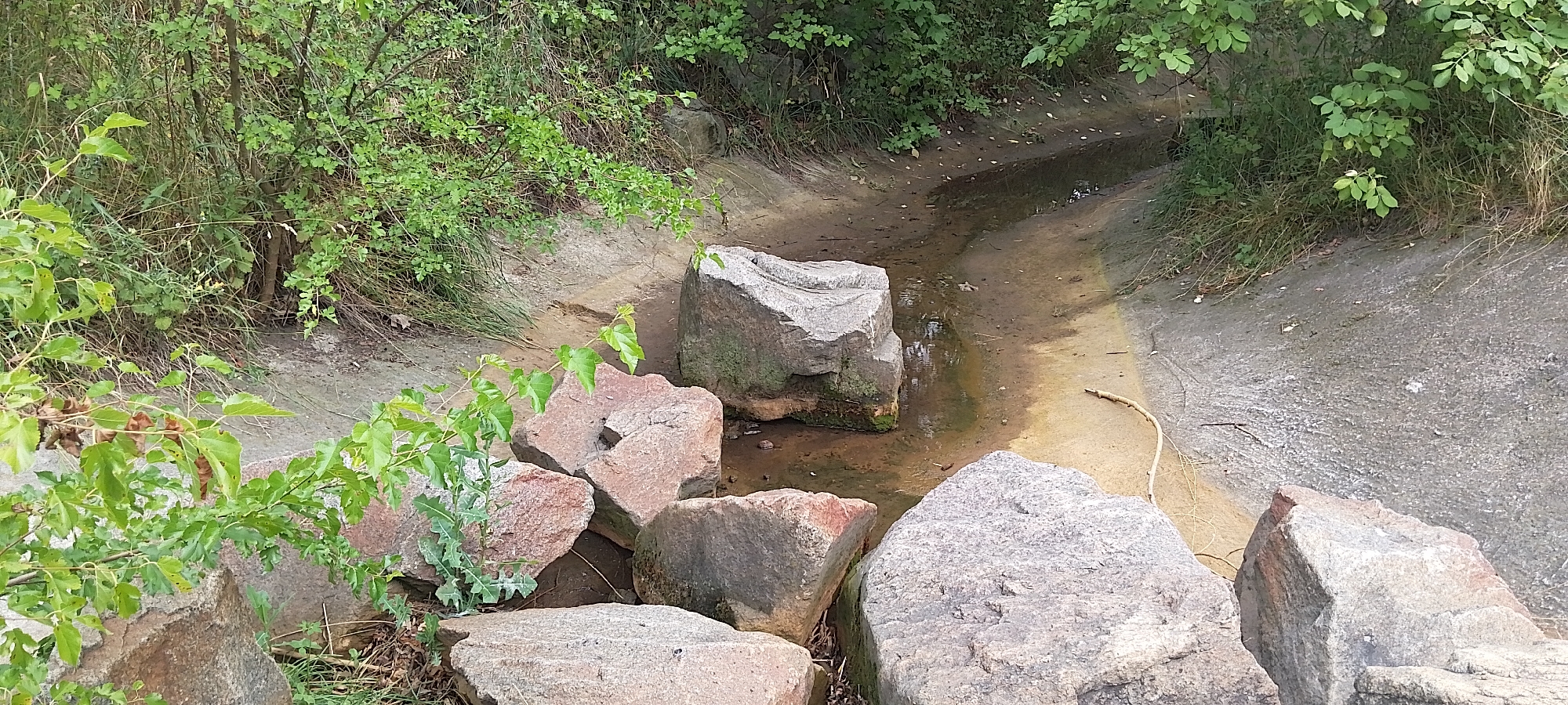 The image depicts a dry or low-water drainage channel in Cherkasy, Ukraine. The channel has concrete sides in some areas and is lined with soil and sand. Several large, rough-hewn rocks are situated in and around the channel, some partially submerged in shallow water. Green foliage, including trees and bushes, borders the channel. The scene appears to be an outdoor, undeveloped area within the city.