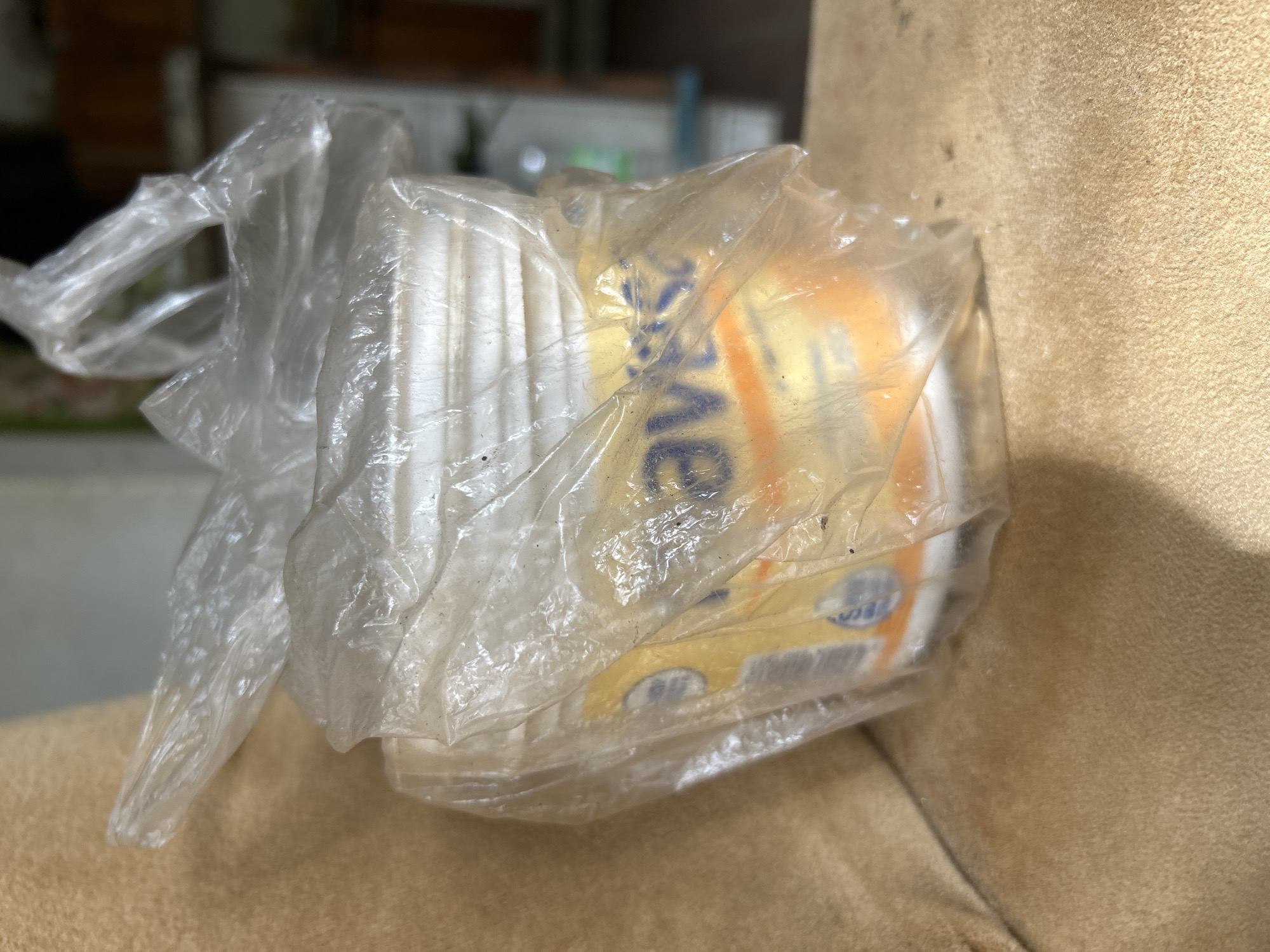A plastic bag containing a white bucket with a yellow label, partially obscured by the bag's plastic, rests on a textured brown surface. The label on the bucket appears to have blue and orange text. The background is blurred, showing hints of shelves and a light-colored object. The scene suggests an indoor setting, possibly a home or storage area, located in Santa Ana, Venezuela.