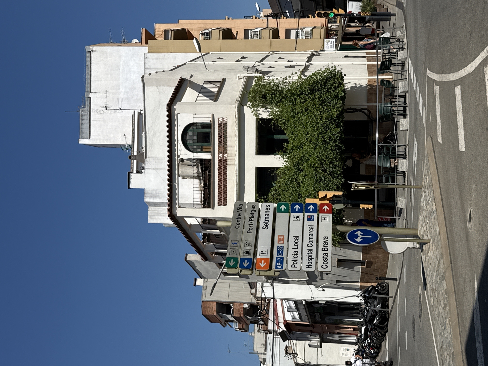 The image captures a street scene in Blanes, Spain, on a clear, sunny day. In the foreground, a pedestrian crossing is visible with white lines painted on the asphalt. A signpost with multiple directional signs is prominently displayed. These signs indicate directions to "Centre Vila," "Port/Platges," "Setmanes," "AP-7 C-32 N-II," "Policia Local," "Hospital Comarcal," and "Costa Brava." A blue circular sign with two white arrows pointing left and right is also present.
Behind the signpost, a white building with red-tiled roof accents stands. It features balconies with decorative railings and a prominent arched window. To the right, a taller, pale orange building is visible, also with balconies. The street in front of these buildings appears to be a town square or intersection. A dense green tree with thick foliage provides shade to an outdoor seating area with tables and chairs.
Several motorcycles are parked in a row on the left side of the image, near the buildings. A few people are present in the scene: a person in a white top and dark bottoms is walking towards the motorcycles, and other individuals are seen further in the background, some near the outdoor seating area and others further down the street. A traffic light with a green light illuminated is visible on the right side of the image. The overall impression is a bustling urban environment on a warm day.
