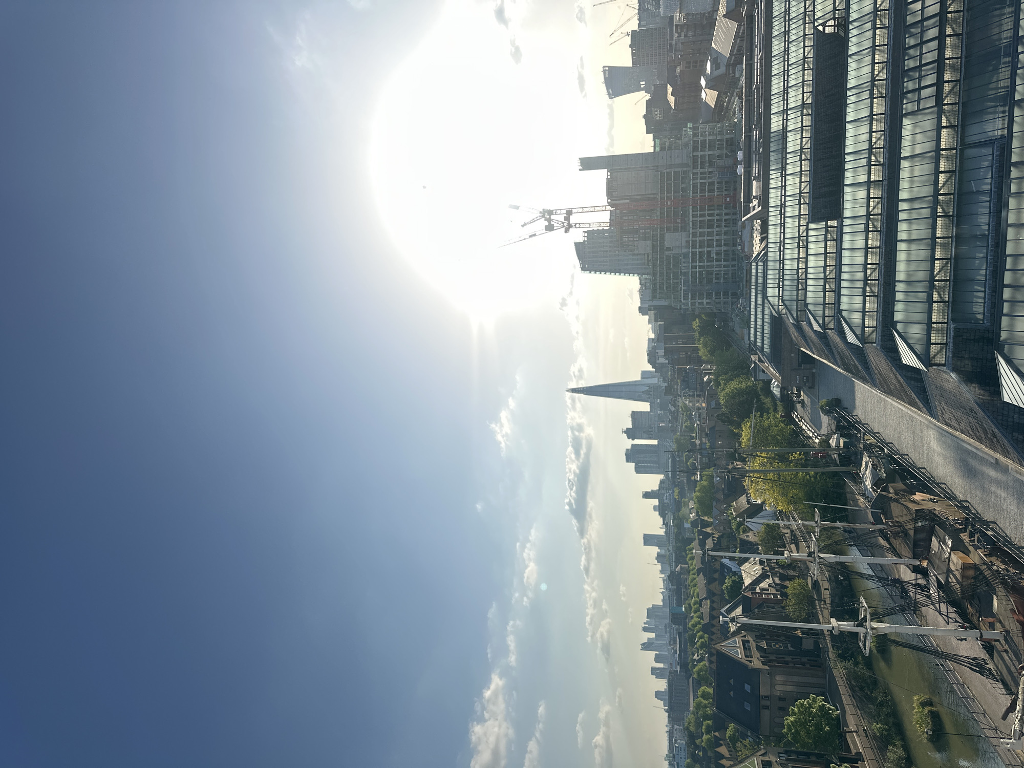 The image presents an expansive, high-angle view of a sun-drenched cityscape in London, United Kingdom, taken on a clear day. The sun, a brilliant, overexposed circle, dominates the upper left portion of the sky, casting a bright glare and lens flare across the scene. The sky transitions from a deep blue on the far right to a lighter, almost white hue near the sun, with a few scattered white clouds.
The urban landscape unfolds below, a blend of historical and modern architecture. In the upper center, the distinctive pointed silhouette of The Shard stands tall, a prominent landmark. To the left, a large contemporary building with an extensive glass and metal roof structure stretches into the frame, likely a commercial or transport hub. Several other modern high-rise buildings, some under active construction with visible cranes, are scattered across the mid-ground, indicating ongoing development.
In contrast, the lower right section of the image reveals a more historic and densely packed area of lower-rise buildings, characterized by traditional roofing and tree-lined streets. A narrow waterway, possibly a canal or a section of the Thames, snakes through this part of the city. Moored within this waterway is a large, multi-masted sailing ship, its intricate rigging clearly visible, adding a historical maritime element to the contemporary urban backdrop. Patches of lush green trees are interspersed among the buildings, providing natural accents.
No specific human activity is visible, but the bright sunlight suggests a vibrant day. The overall impression is one of a bustling metropolis, dynamic and evolving, set against a bright, clear sky. There is no discernible text within the image.
