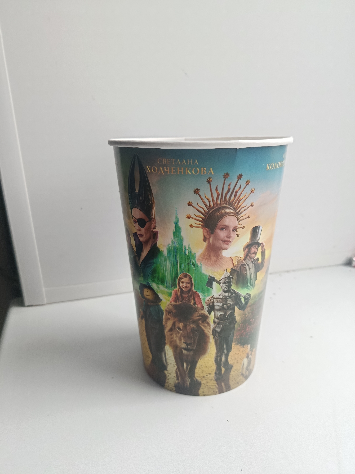 The image shows a close-up, eye-level, medium shot of a paper cup with a colorful illustration on its side, set against a plain white background. The cup is positioned in the foreground, slightly angled to the right, and its bottom edge is out of frame. The illustration features characters from "The Wizard of Oz," including a woman in a tall, horned black headdress and an eyepatch, a woman with a sun-like headdress, a man in a top hat, a tin soldier, a lion, and a young girl. The background of the illustration depicts a green castle and a colorful sky. The cup's white rim is clearly visible. The white background suggests an indoor setting, likely a table or shelf. There is no indication of the city of Syzran, Russia, in the image.