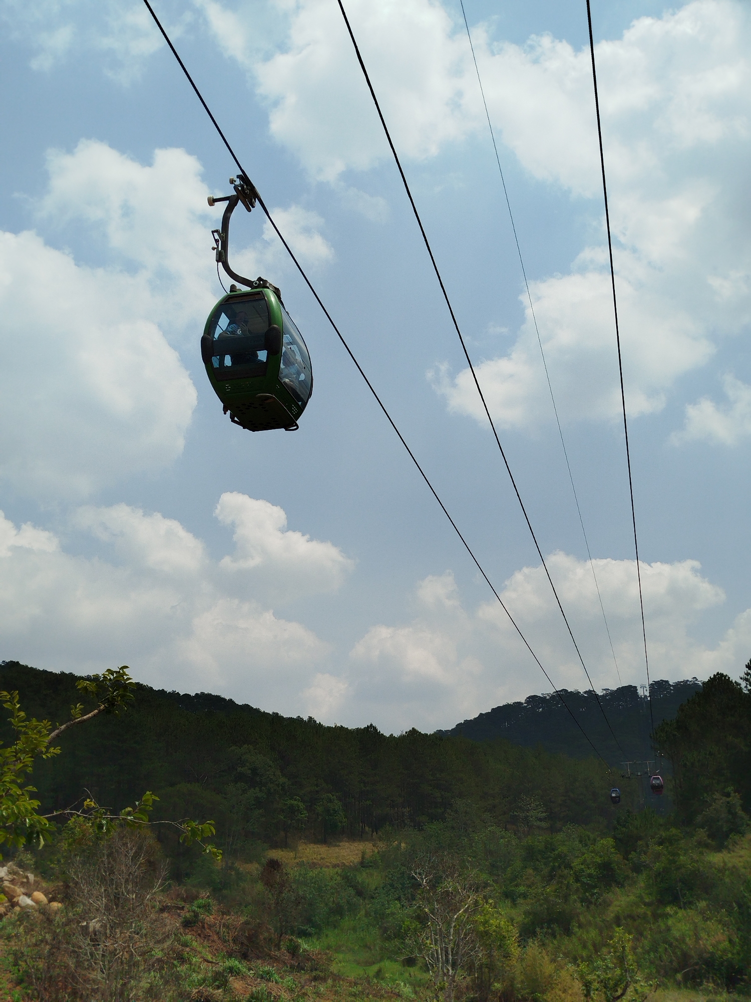 The image depicts a cable car system in Da Lat, Vietnam. A green cable car with visible occupants hangs from multiple cables stretching across the frame. Below, a dense forest covers rolling hills. The sky is partially cloudy with patches of blue. In the distance, another cable car can be seen. The vegetation in the foreground appears to be a mix of trees and scrub brush.