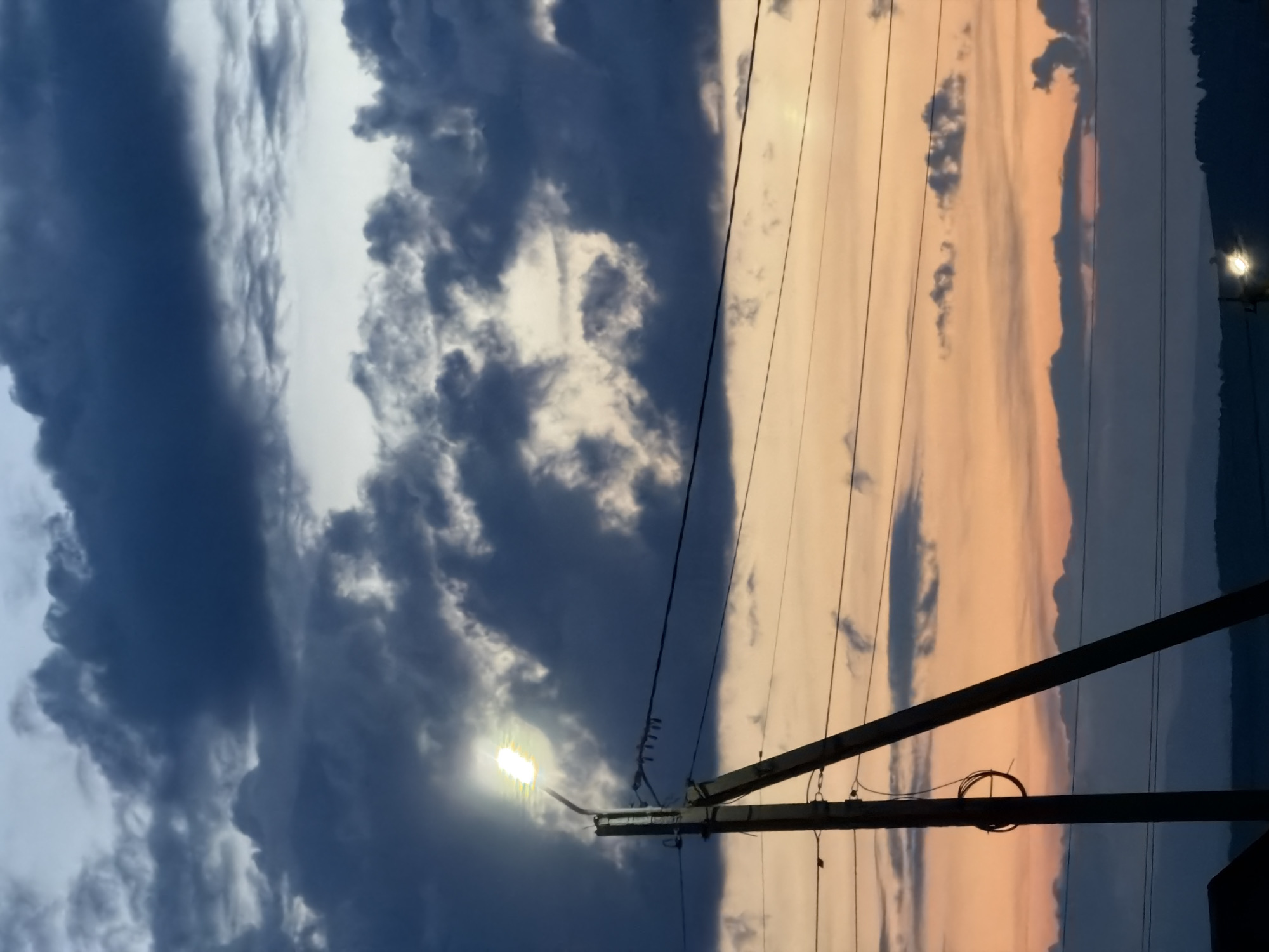 A street lamp illuminates a dusky sky filled with dark, heavy clouds. The lamp, positioned on a tall pole, casts a bright, white light that is partially obscured by the low-hanging clouds. Several power lines are strung between poles, crisscrossing the frame against the backdrop of the twilight sky. The sky transitions from a soft orange near the horizon to a deep blue and gray at the top, indicating either dawn or dusk. In the distance, a dark silhouette of hills or mountains can be seen. Another distant light is visible near the bottom of the frame, possibly another street lamp or a house light. The scene suggests an outdoor, possibly rural or suburban, setting at the beginning or end of the day.