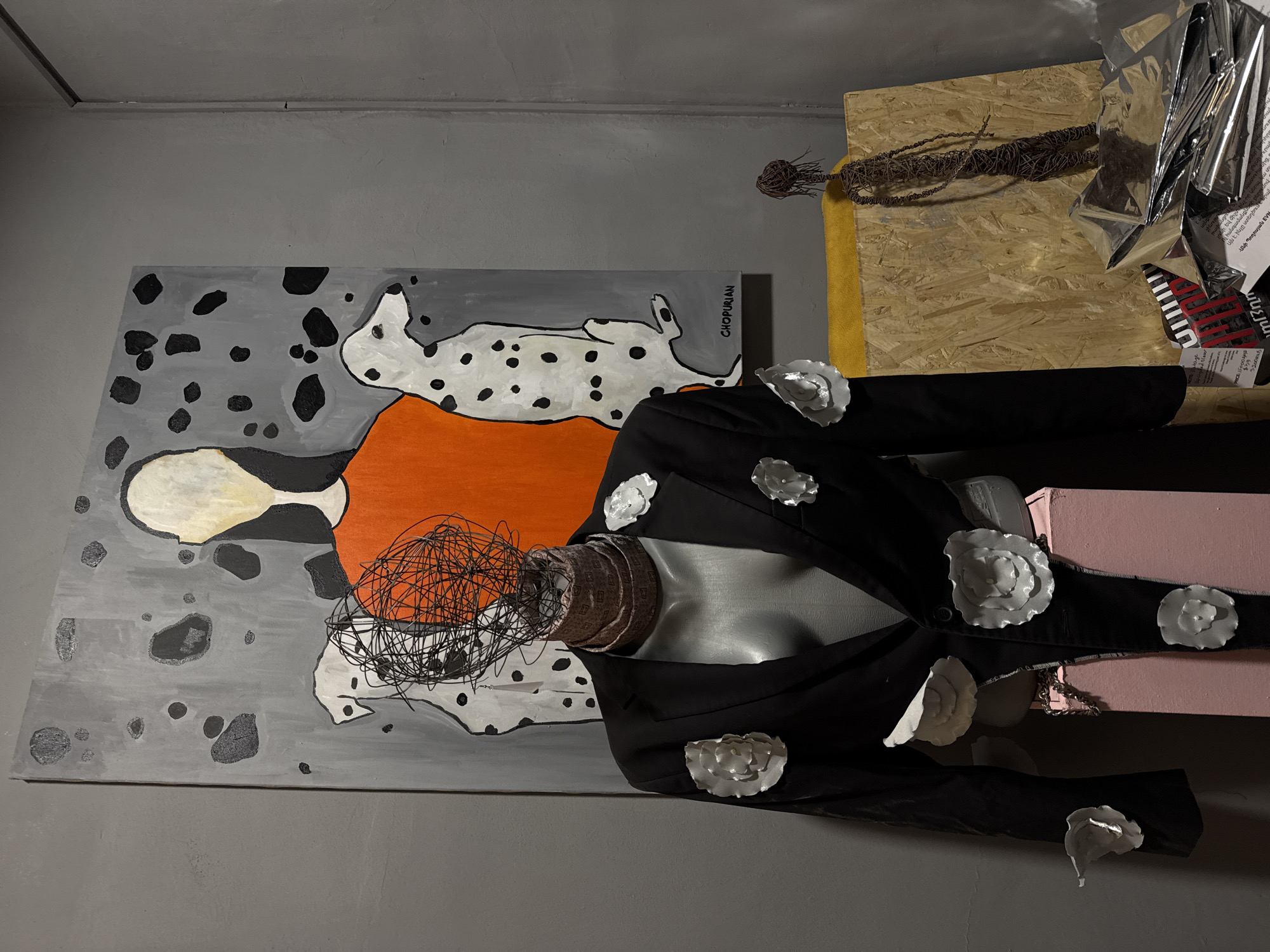 The image captures an artistic installation or display in what appears to be an indoor setting. The scene includes a painting, a mannequin, and various decorative elements. The painting features abstract figures and shapes in a gray, orange, white, and black palette. The mannequin is dressed in a black jacket adorned with white, flower-like embellishments. The mannequin's head is obscured by a tangled wire-like structure. A patterned neck scarf is wrapped around the mannequin's neck. The mannequin rests on pink boxes. In the background, there is a wooden board, some metallic material, and what appears to be dried vines. Text is faintly visible on a piece of paper near the metallic material, indicating the location as Yerevan, Armenia, and a name "Ani Poghosyan EVN." The overall impression is that of an art exhibition or a creative studio.