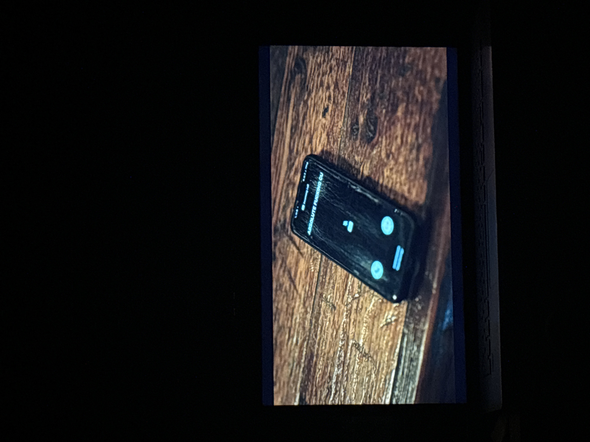 The media file captures a rectangular, brightly lit image, likely a screen or projection, set against a predominantly black background, suggesting a dark viewing environment. The depicted scene features a black smartphone resting diagonally on a wooden surface.
The wooden surface exhibits prominent, warm-toned wood grain, suggesting it could be a table or floor. The smartphone's screen is illuminated and appears to be heavily cracked or scratched. On the screen, light blue (cyan) interface elements are visible, including what looks like control buttons or icons. The most prominent visible text on the phone's display is "ABSOLUTE FUCKING CU", partially cut off at the left edge of the screen. Other circular icons and a vertical bar icon, all in light blue, are also present, hinting at a media playback interface or a communication application.
No individuals are visible in the image. To the right edge of the wooden surface, a dark, textured object, possibly a book spine, can be partially seen. The scene depicts a stationary phone in an indoor setting, with the only illumination coming from the device's screen and the display it is on. There are no direct visual cues within the image itself to specifically indicate the location as Budva, Montenegro, for the scene shown on the screen.