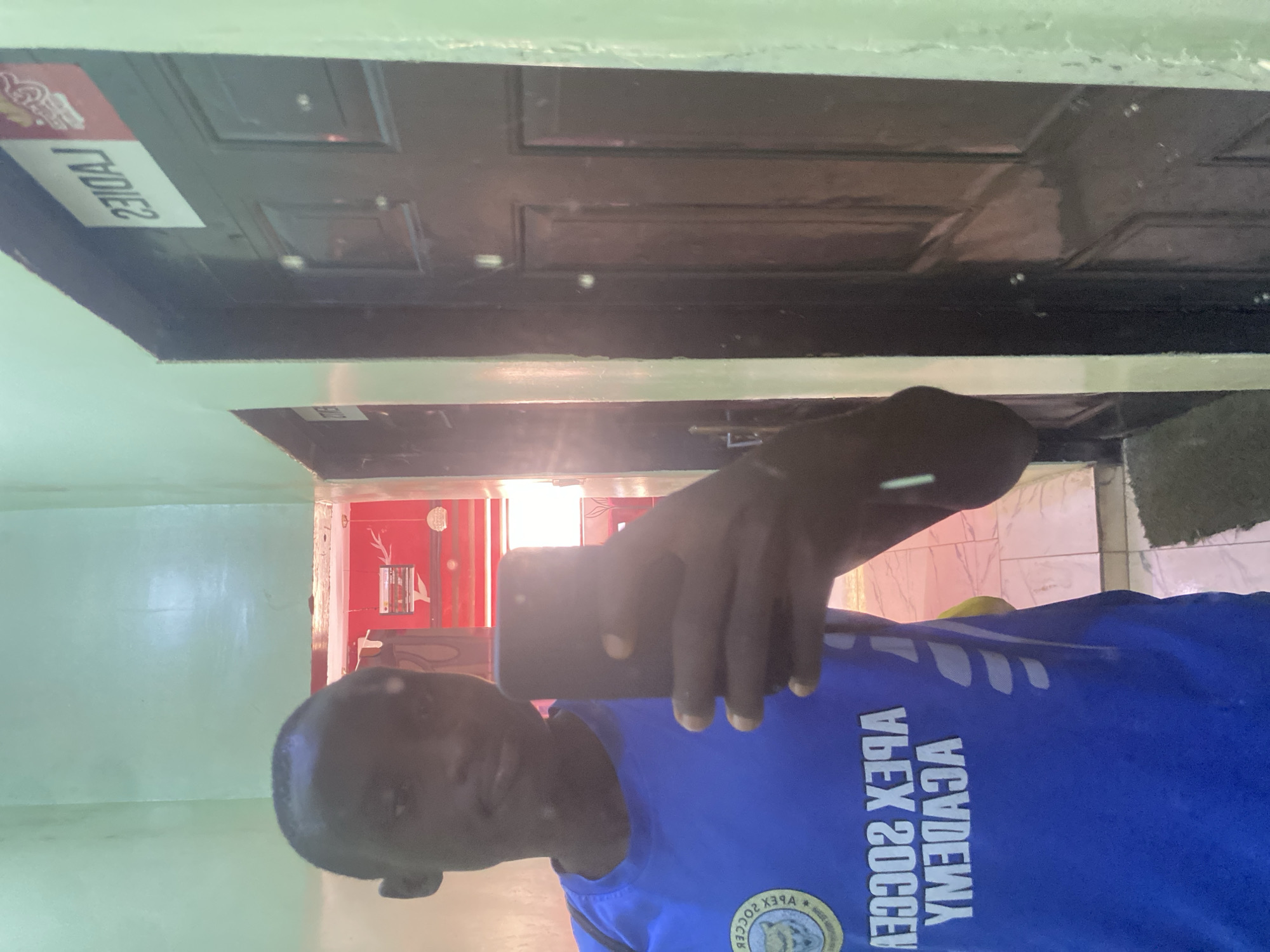 A selfie photograph shows a man wearing a blue soccer jersey with "Apex Soccer Academy" written on it. The photo seems to be taken indoors, with a door labeled "Ladies" visible in the background. The room appears to be simple, with green walls. While the location is reported as Tukur Tukur, Nigeria, there are no specific elements in the image that would verify this geographic information.